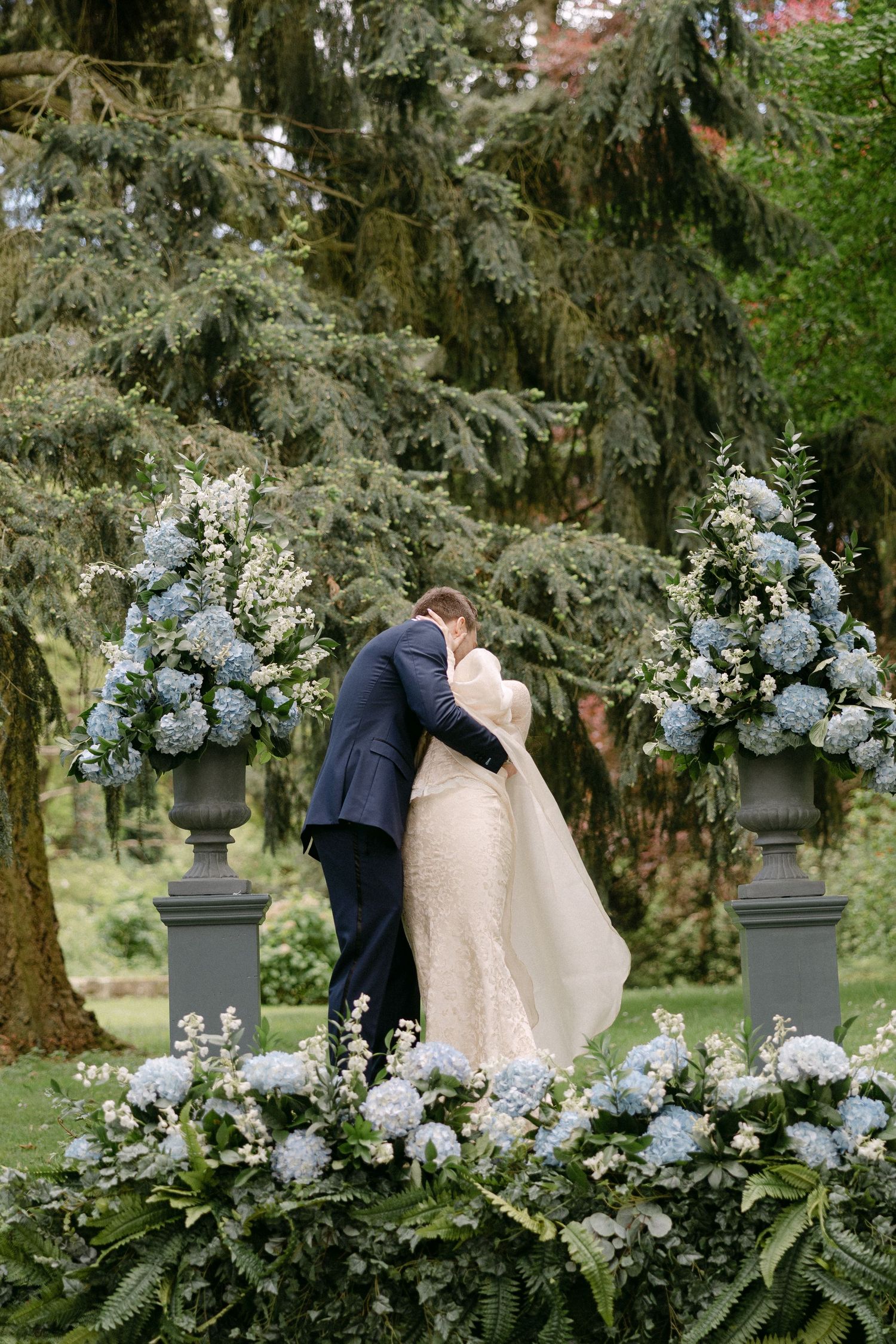 Bride and groom share first kiss at outdoor ceremony surrounded by blue hydrangea floral arrangements.