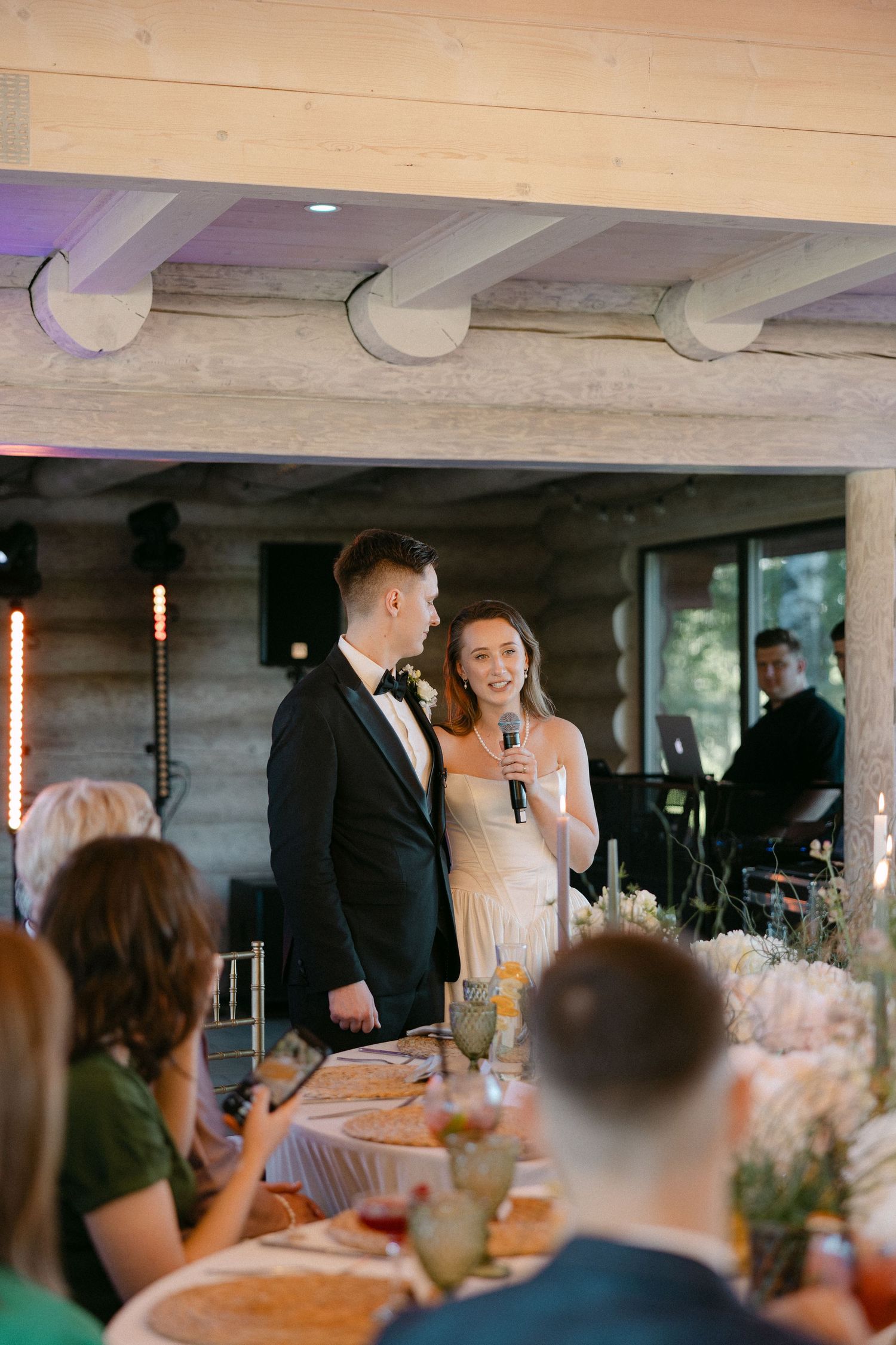 Smiling bride speaks into microphone beside groom during wedding reception speech at decorated banquet table.