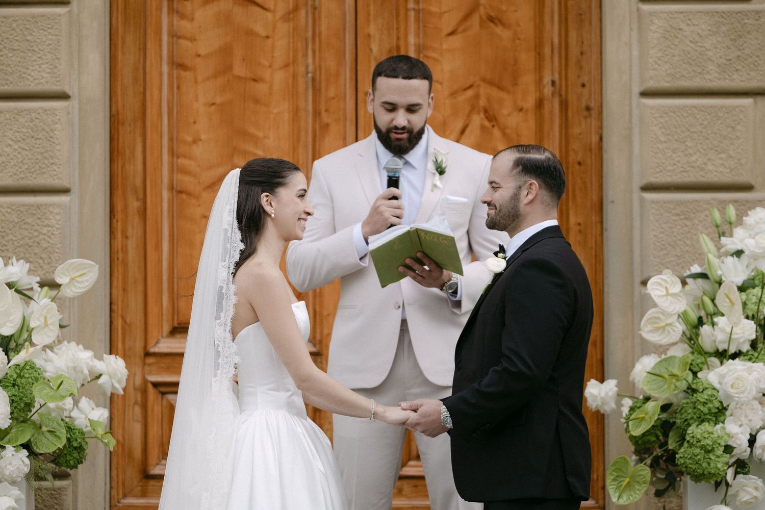 Bride and groom holding hands during wedding ceremony while officiant reads vows at floral-decorated altar.