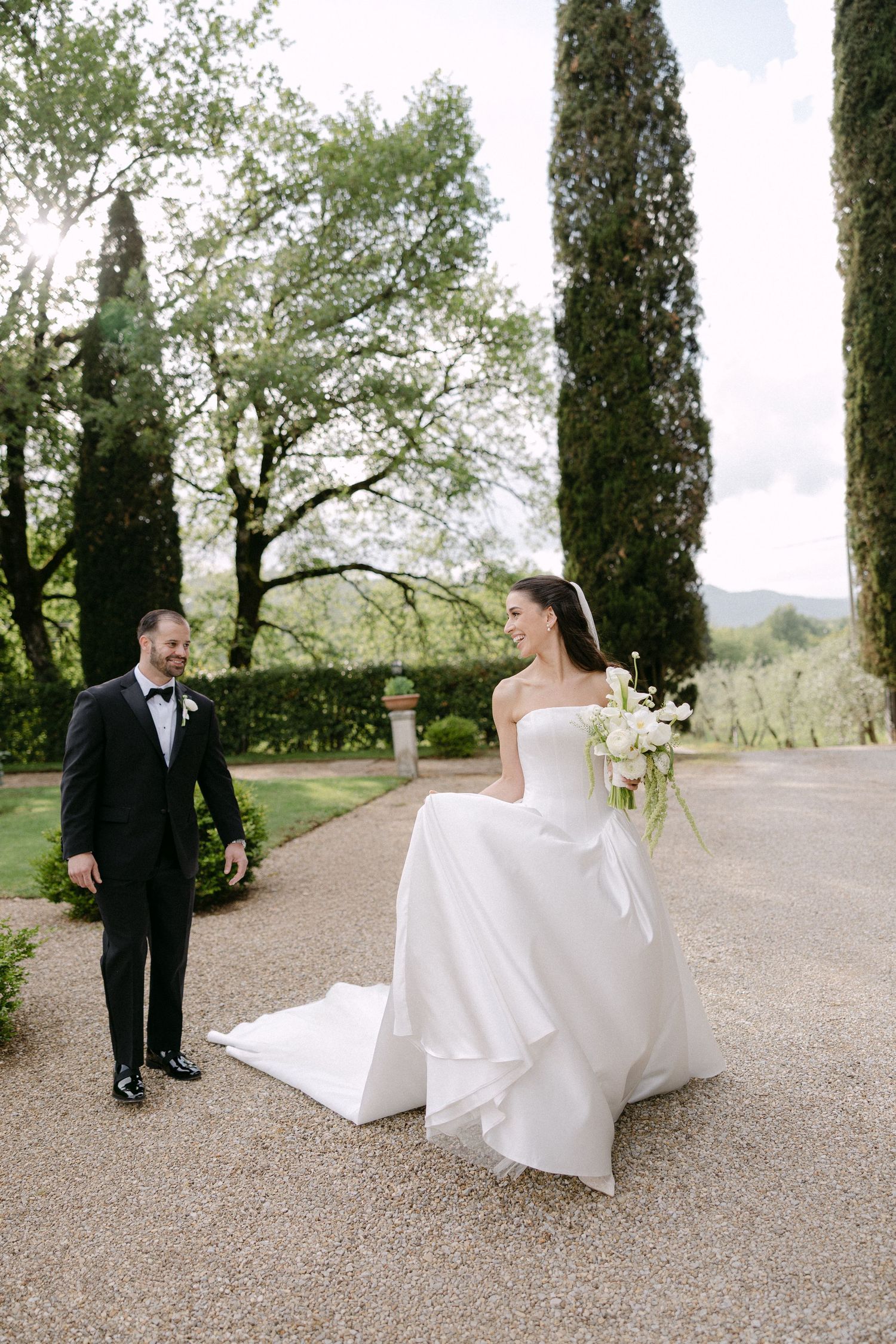 Bride in strapless white gown holds bouquet as groom in black tuxedo walks toward her in lush garden.