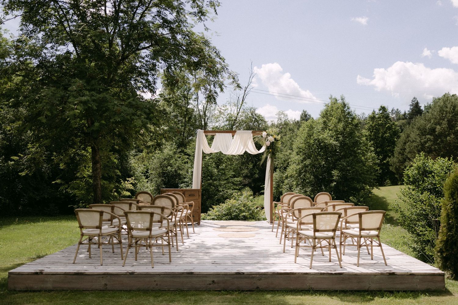 Outdoor wedding ceremony setup with wooden chairs, white arch, and lush green trees on sunny day.