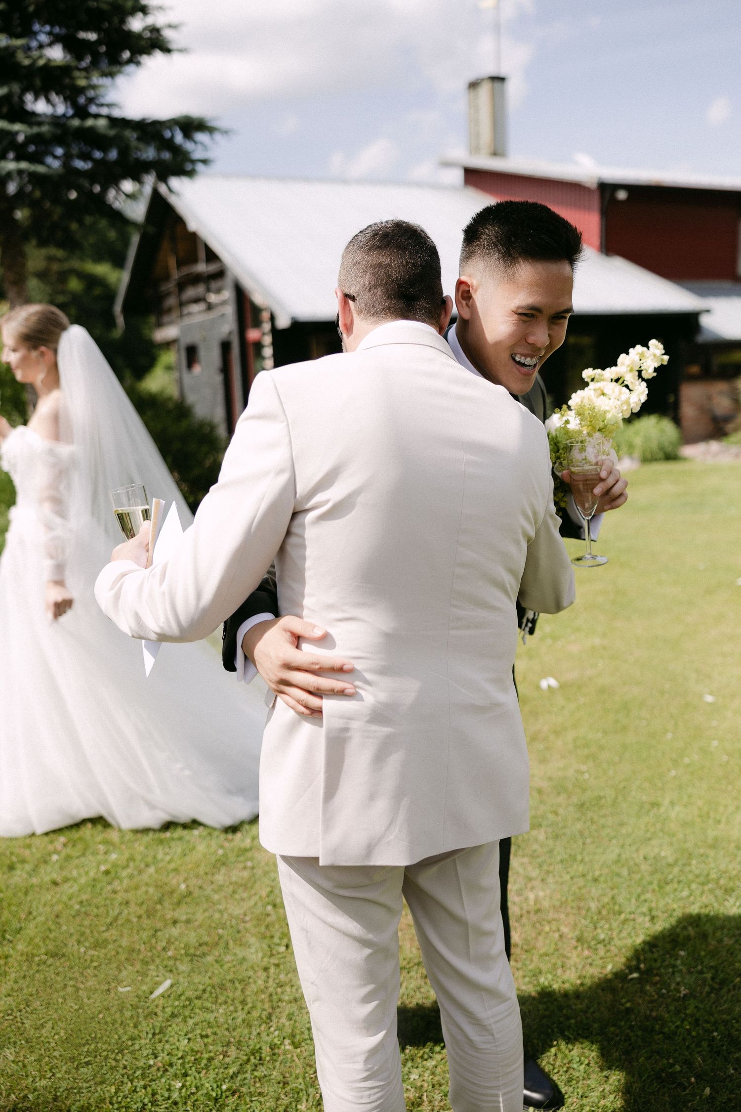 Groom in white suit hugs smiling guest holding flowers at outdoor wedding reception on green lawn.