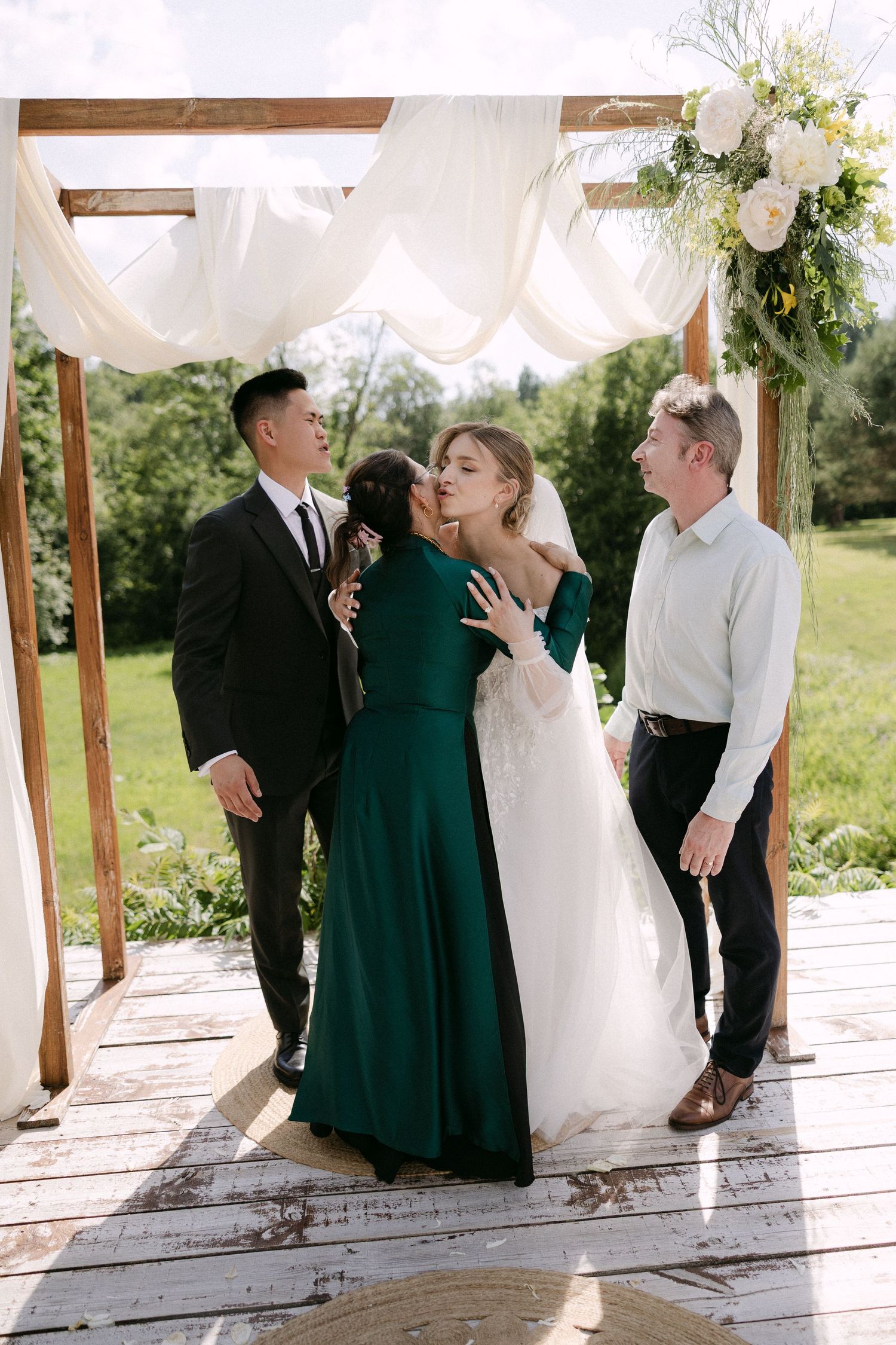 Bride receiving a kiss from a guest in a green dress under a wooden arch at an outdoor wedding ceremony.