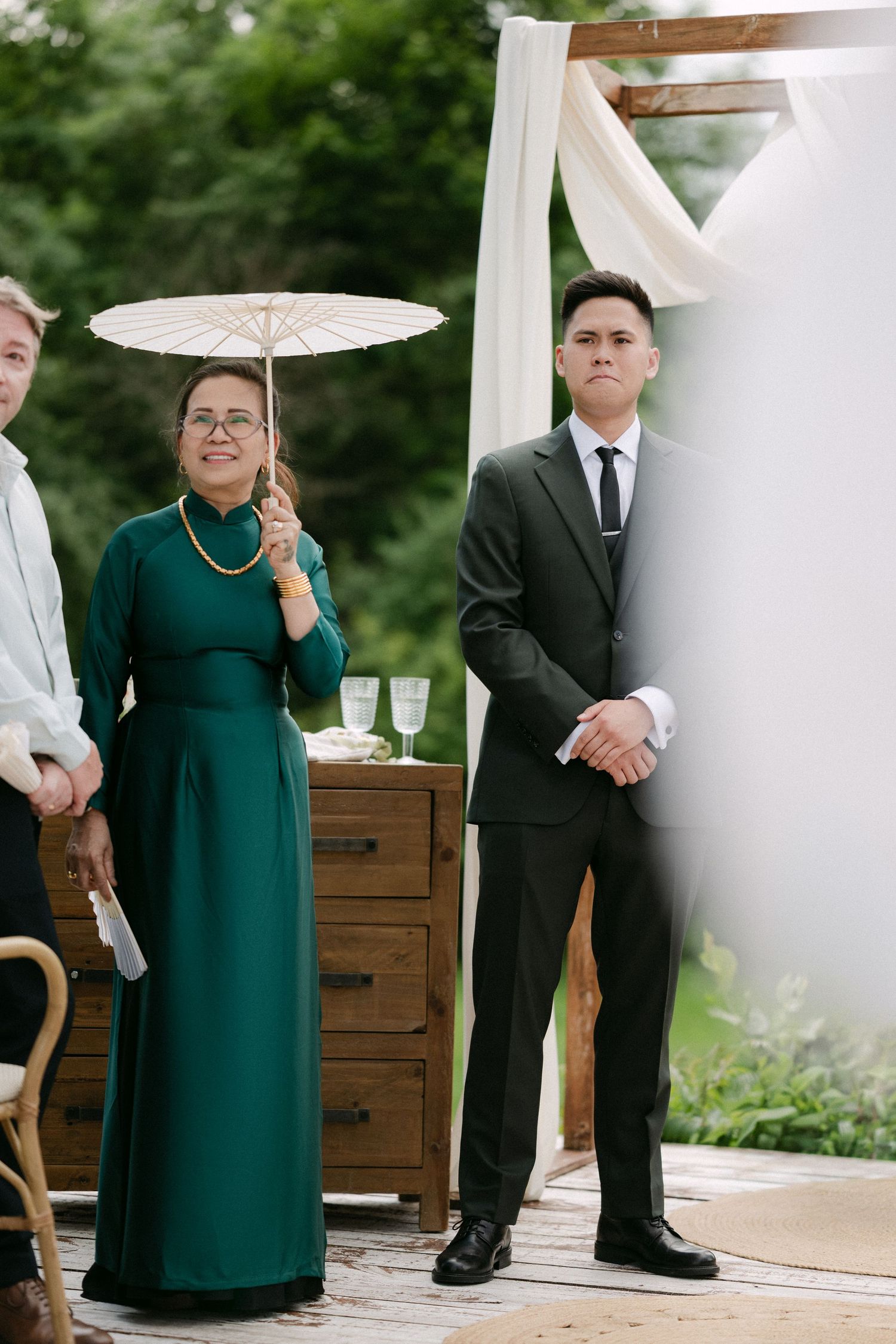 Groom in dark suit stands at outdoor wedding altar beside woman in green ao dai holding white parasol.