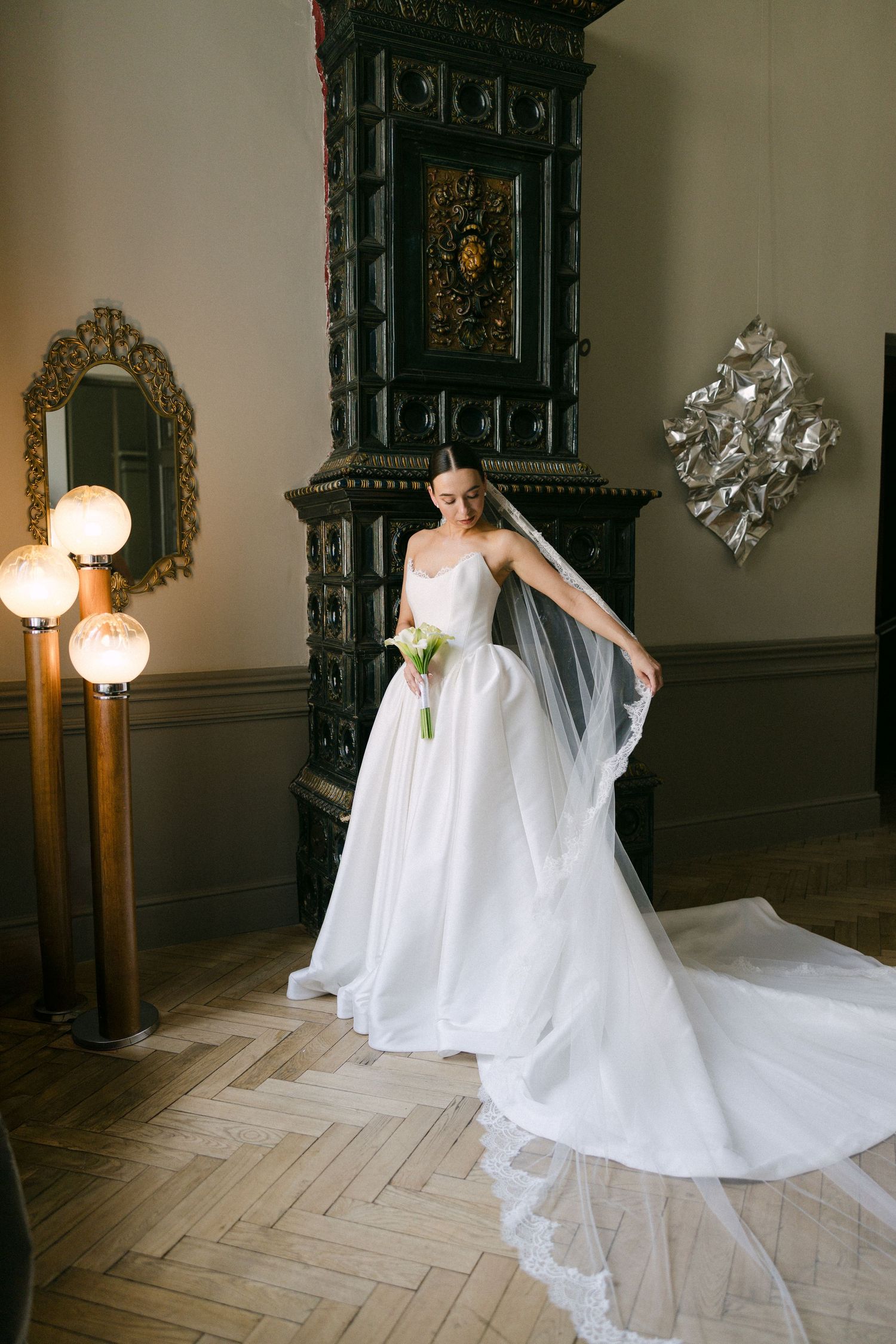Bride in a strapless white ball gown with veil poses before ornate dark fireplace on herringbone floor.