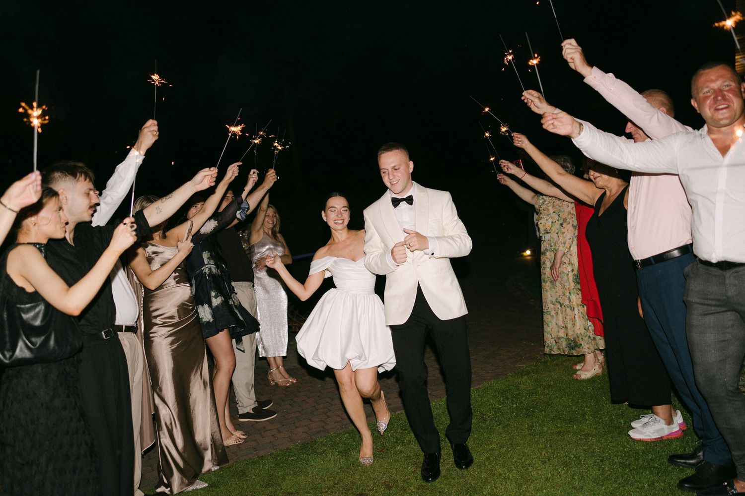 Bride and groom run through a sparkler tunnel surrounded by cheering wedding guests at a night celebration.