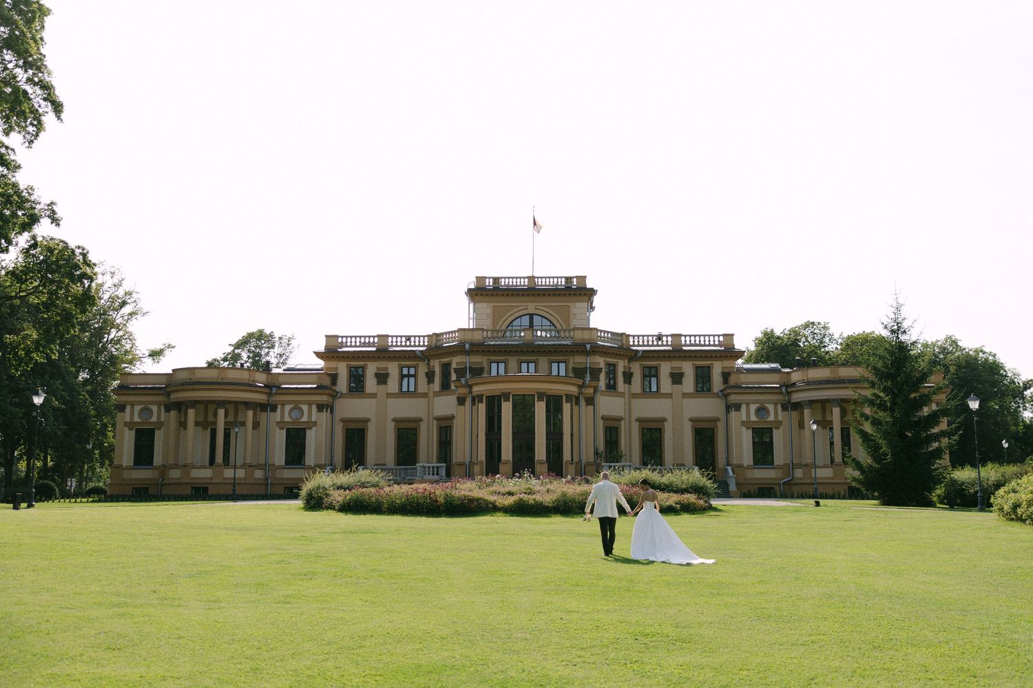 Bride and groom walking hand in hand on lush green lawn in front of elegant historic mansion wedding venue.