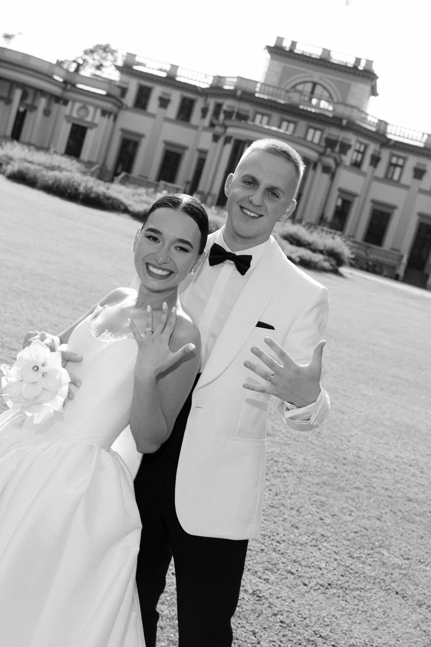 Black and white photo of smiling newlyweds showing wedding rings before grand estate building.