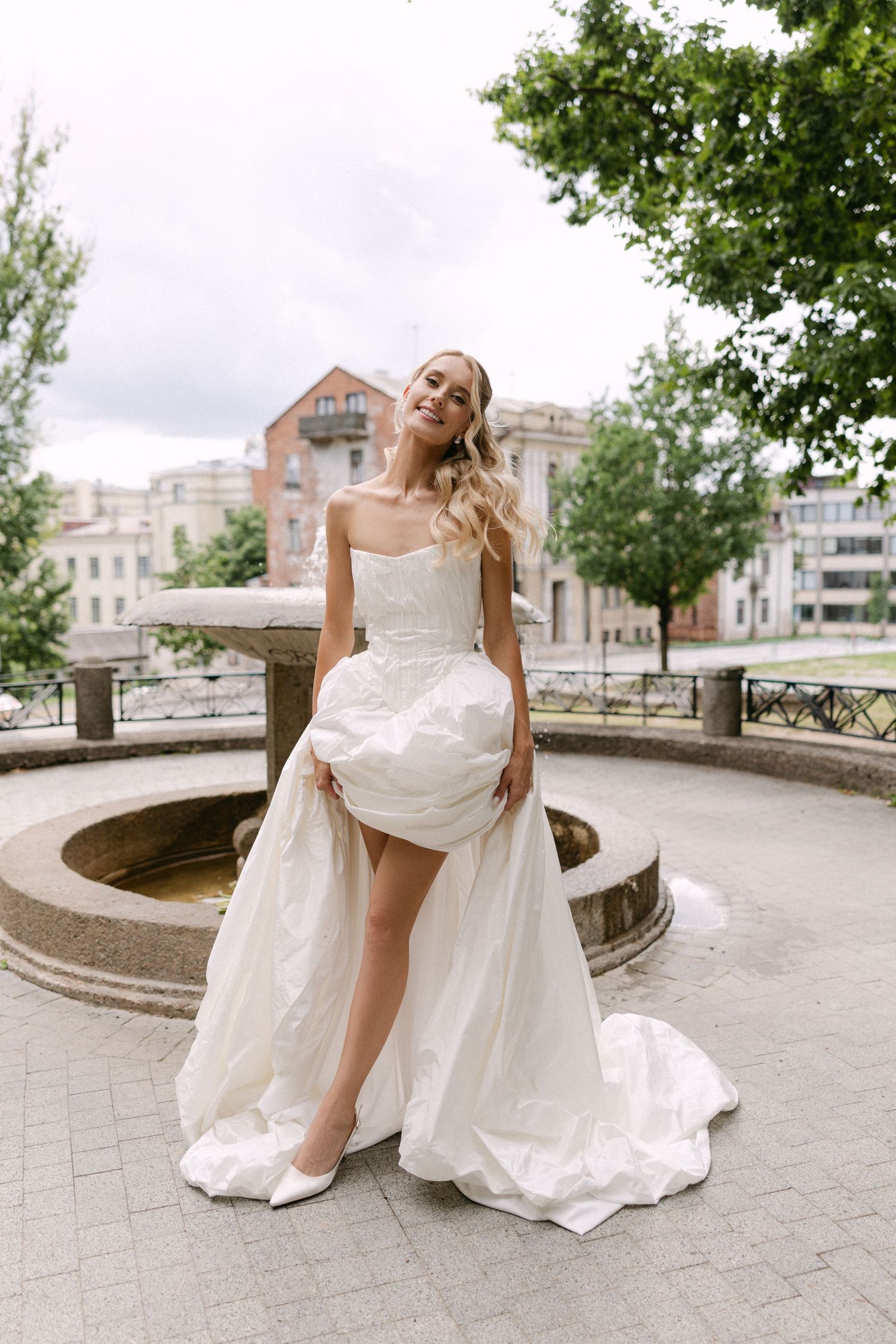 Elegant bride in strapless white wedding gown lifting skirt near stone fountain in urban setting.