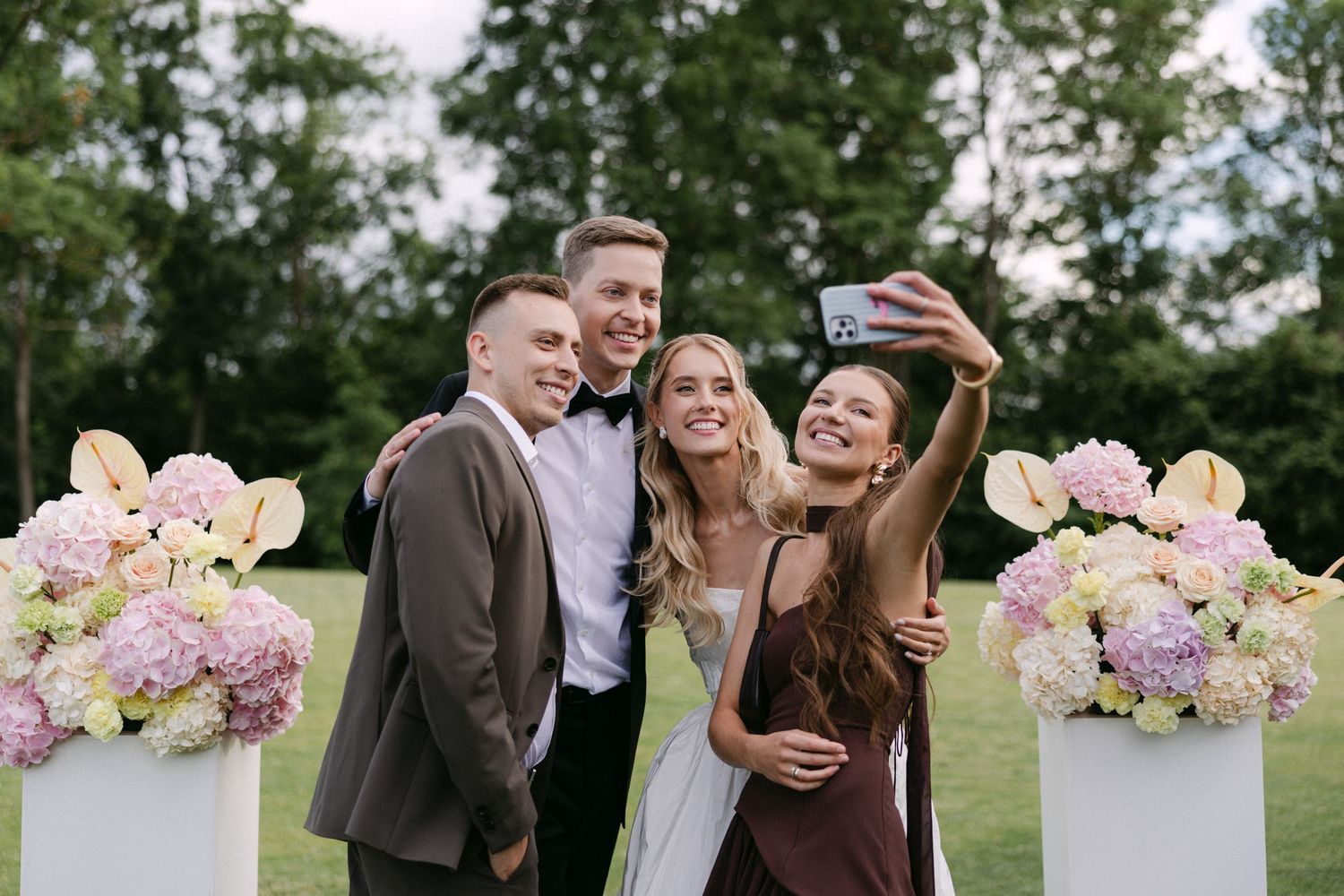 Bride, groom, and two guests taking a fun selfie beside floral arrangements at an outdoor wedding reception.