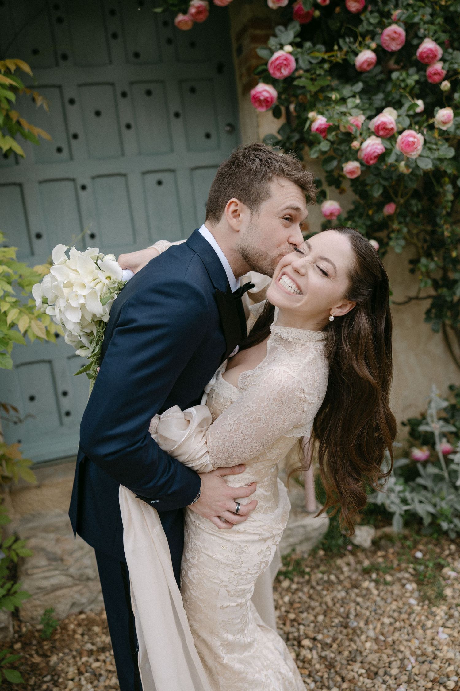 Groom kissing bride on cheek as she laughs joyfully beneath a blooming pink rose-covered doorway.