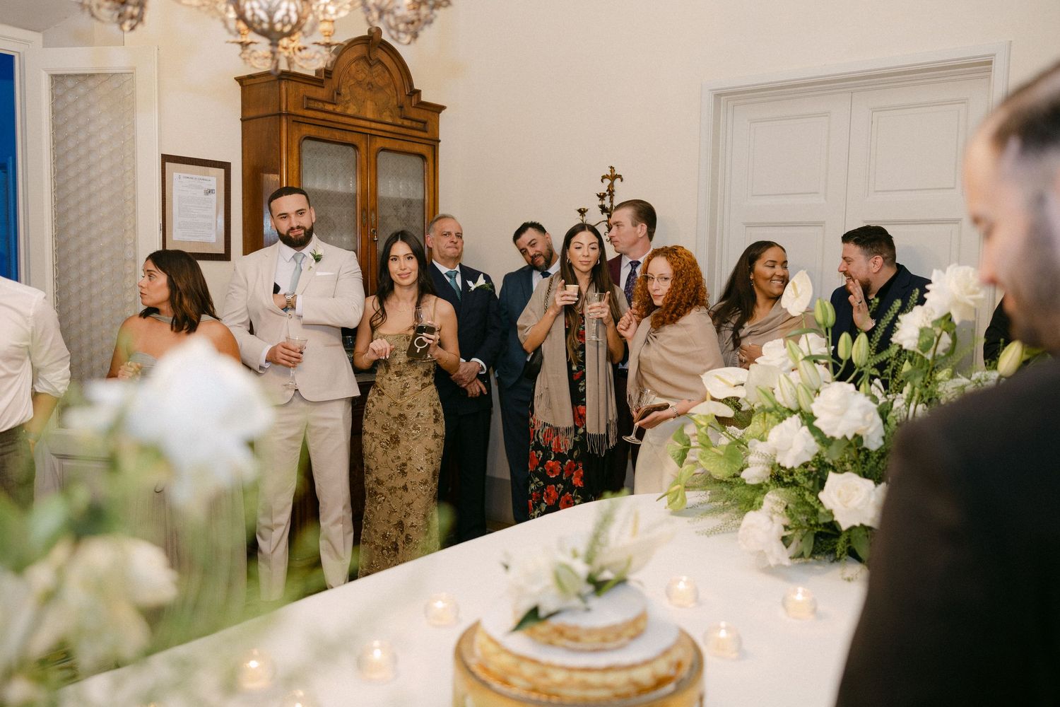 Wedding guests raise glasses in a toast inside elegant white reception room with floral centerpieces.