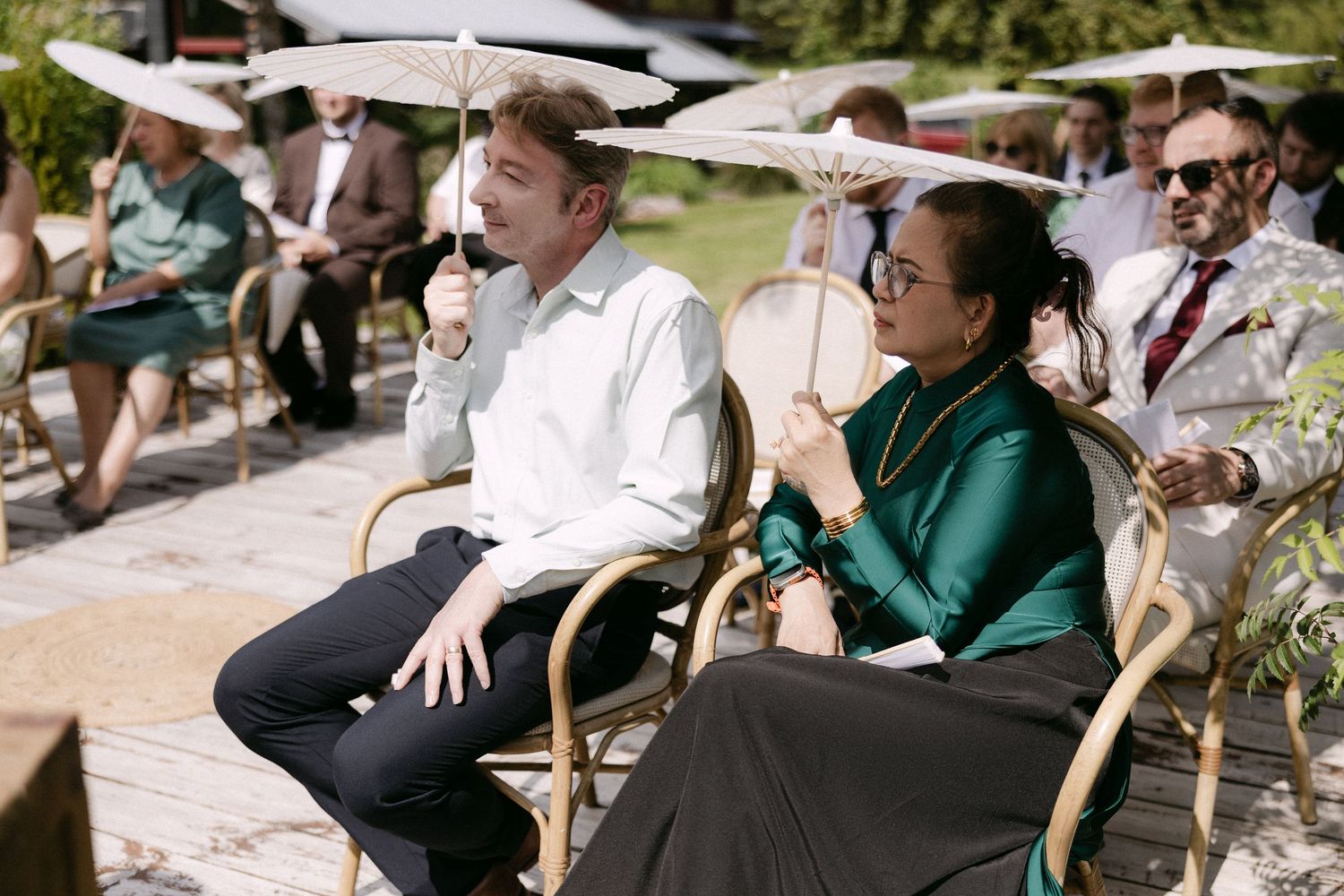 Wedding guests seated in rattan chairs holding white parasols during an outdoor garden ceremony.