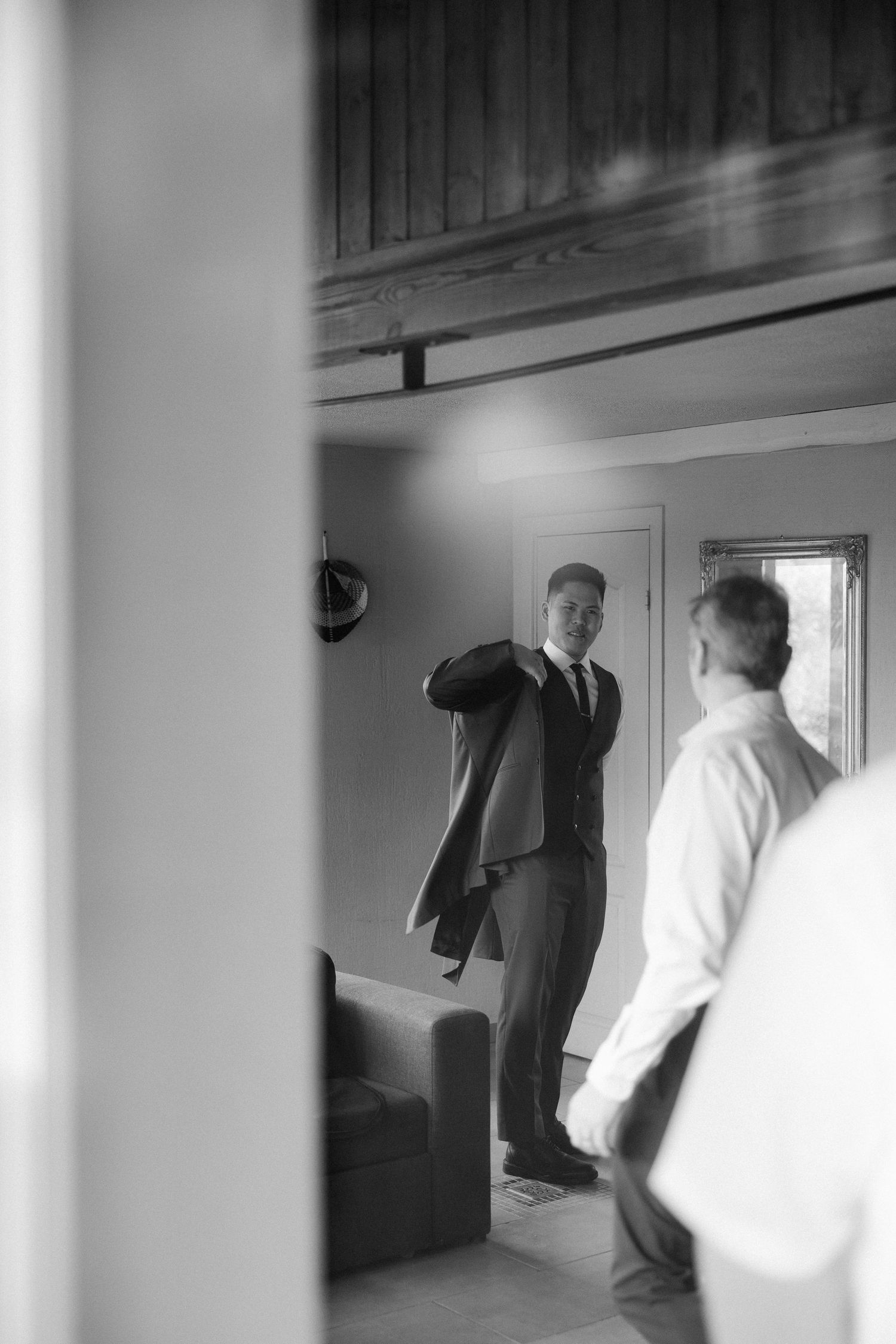 Groom putting on suit jacket in black and white photo while groomsman watches in rustic venue room.