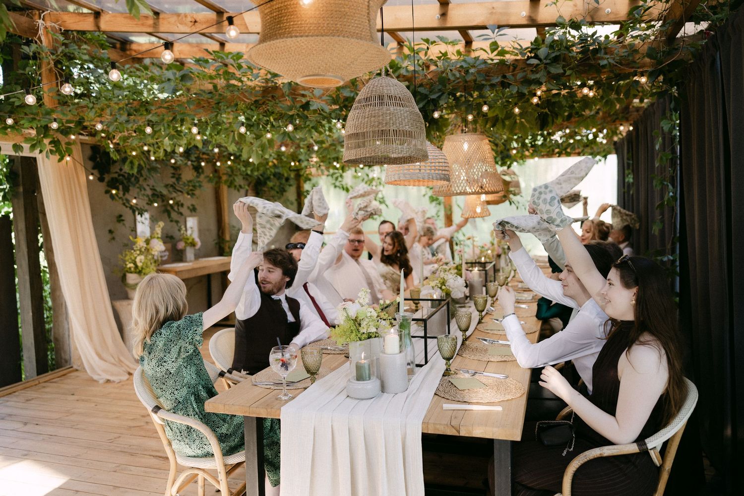 Wedding guests celebrate at long decorated table under pergola with string lights.