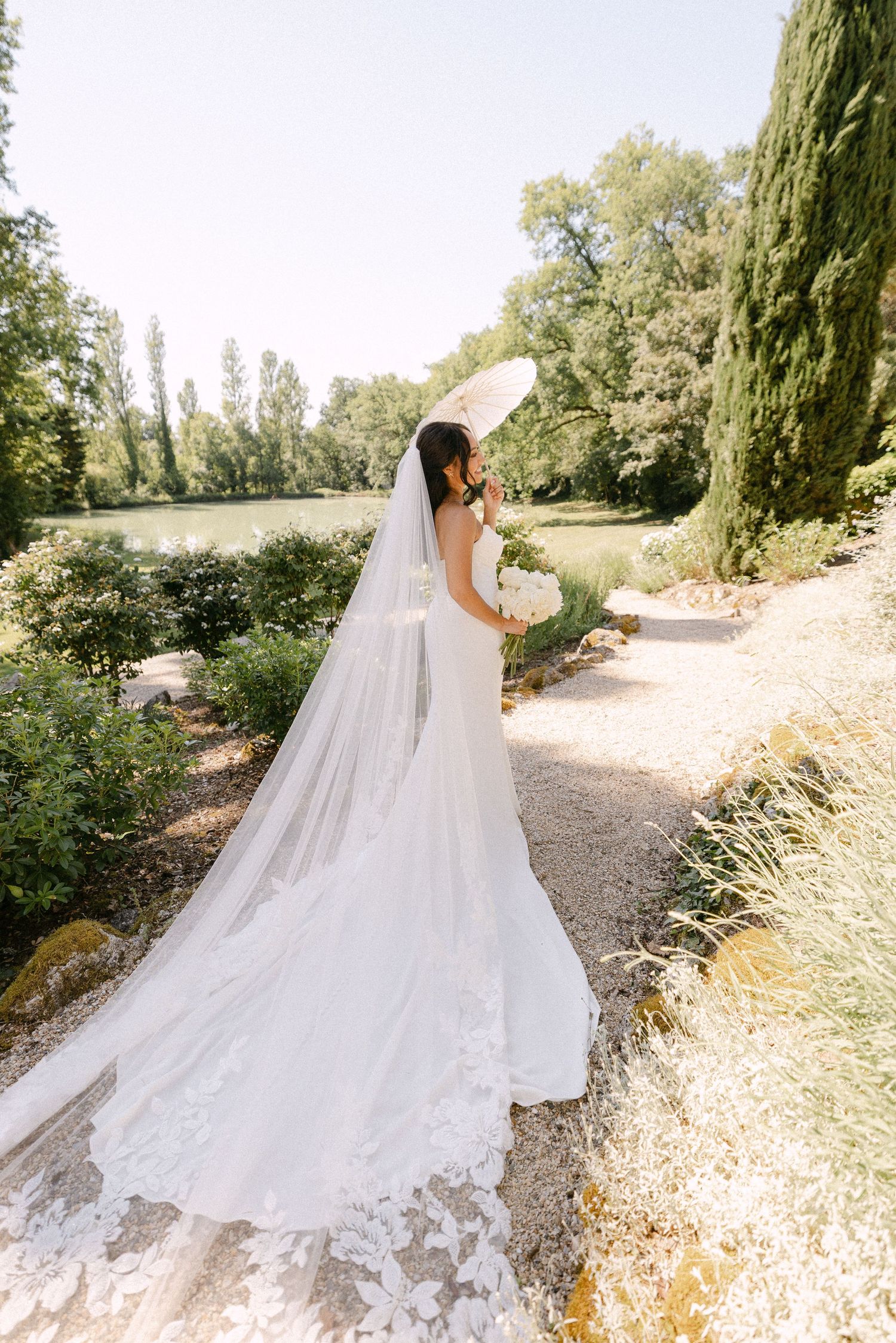 Bride in a stunning white lace wedding gown with a long floral veil holding a bouquet in a lush garden.