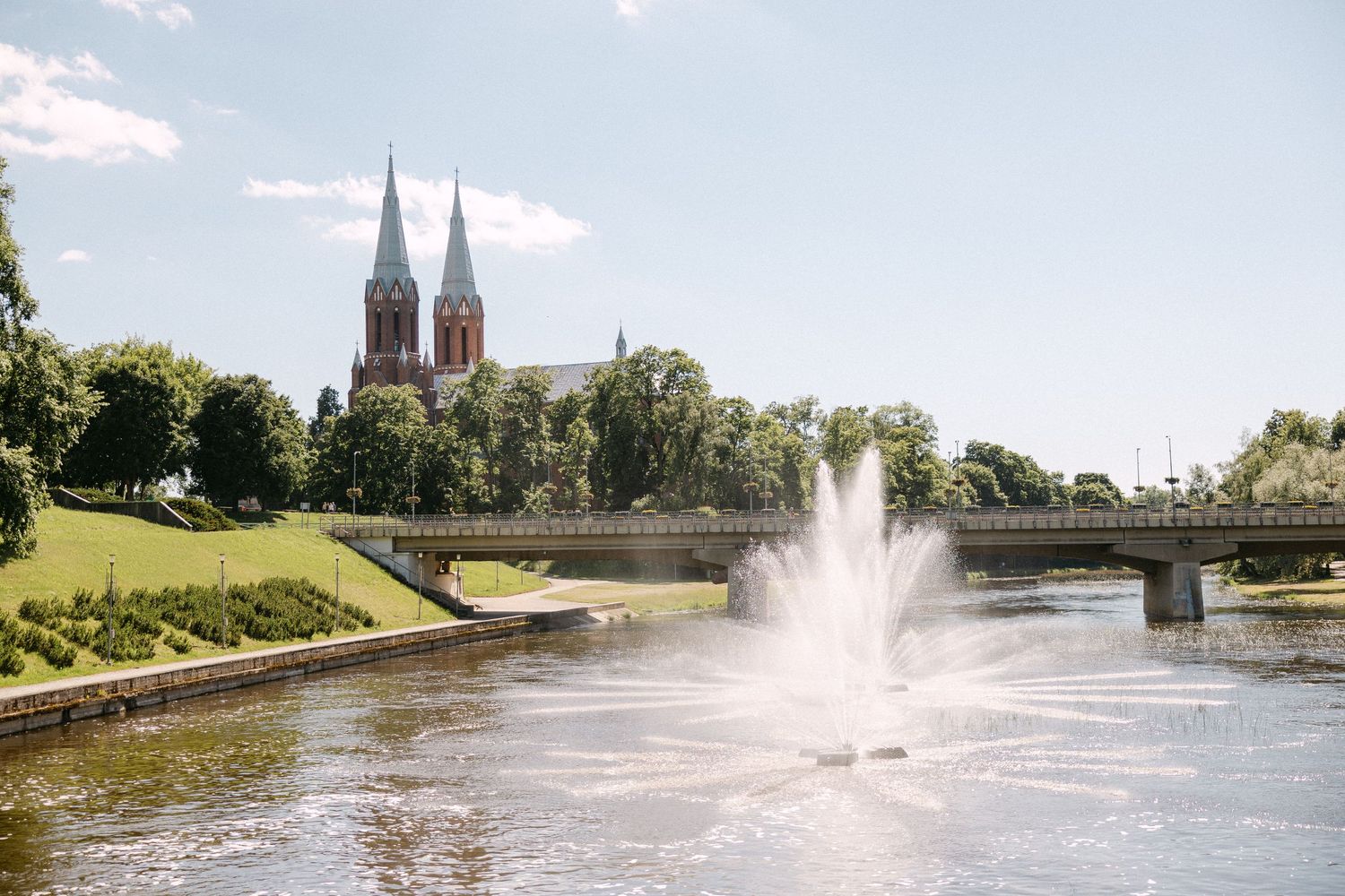 A fountain sprays water on a river with a bridge and Gothic church twin spires rising above lush green trees.