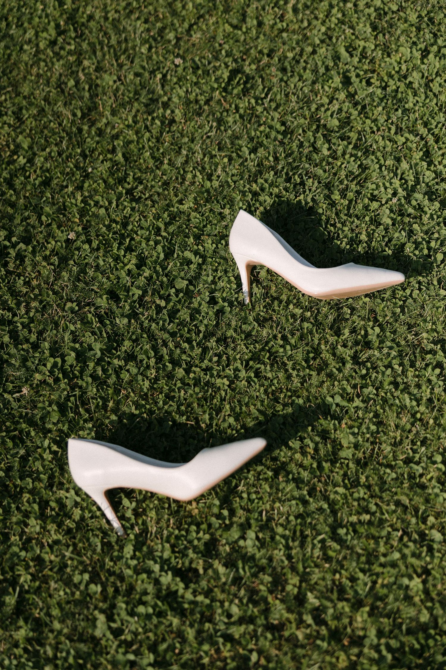 Elegant white bridal high heel shoes placed on lush green grass in natural sunlight.