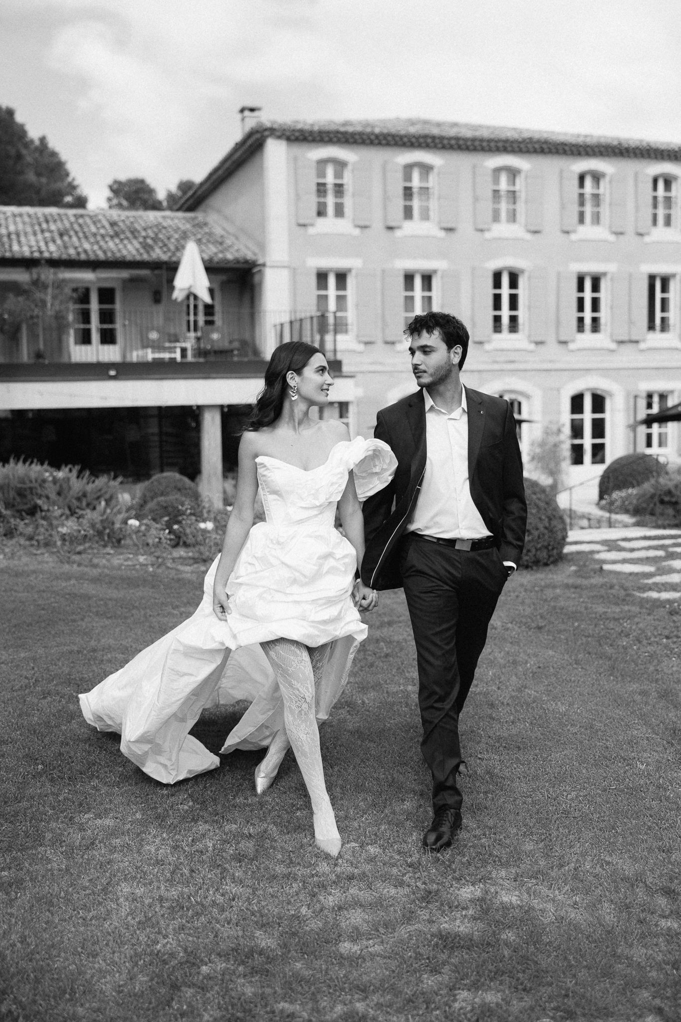 Black and white photo of a bride and groom walking hand in hand on a lawn in front of an elegant estate.