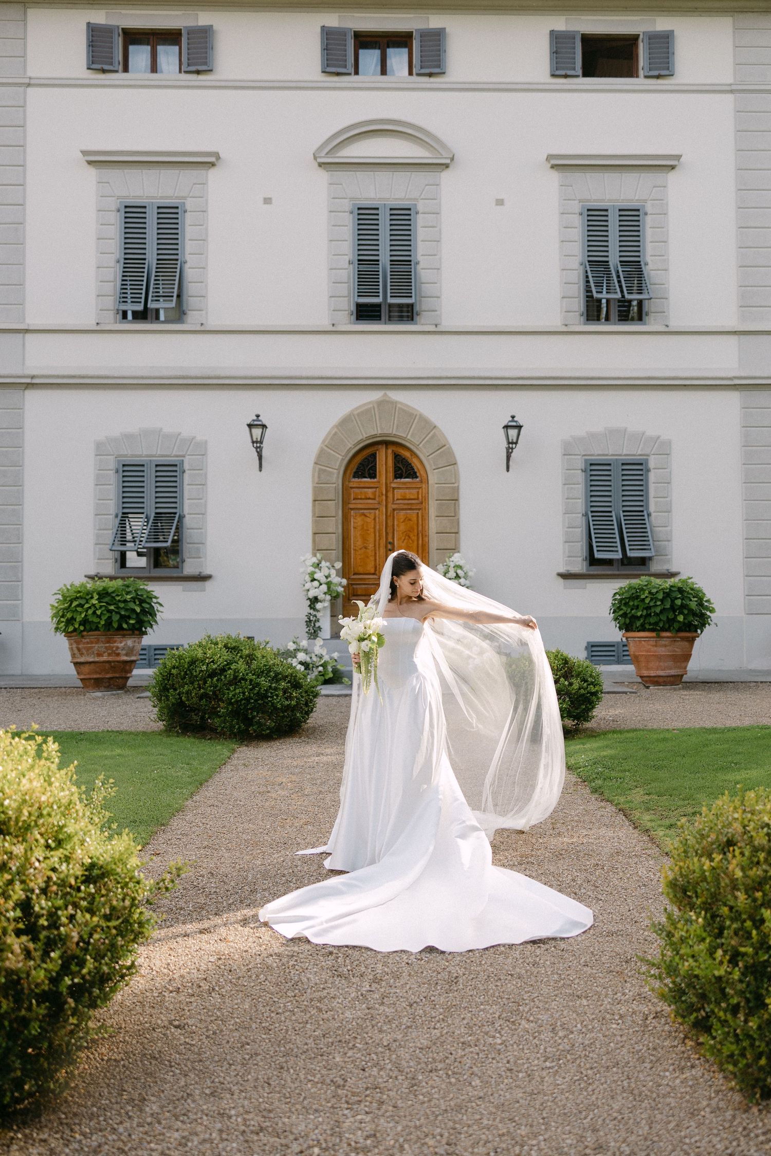 Bride in flowing white gown and veil posing before elegant Italian villa.