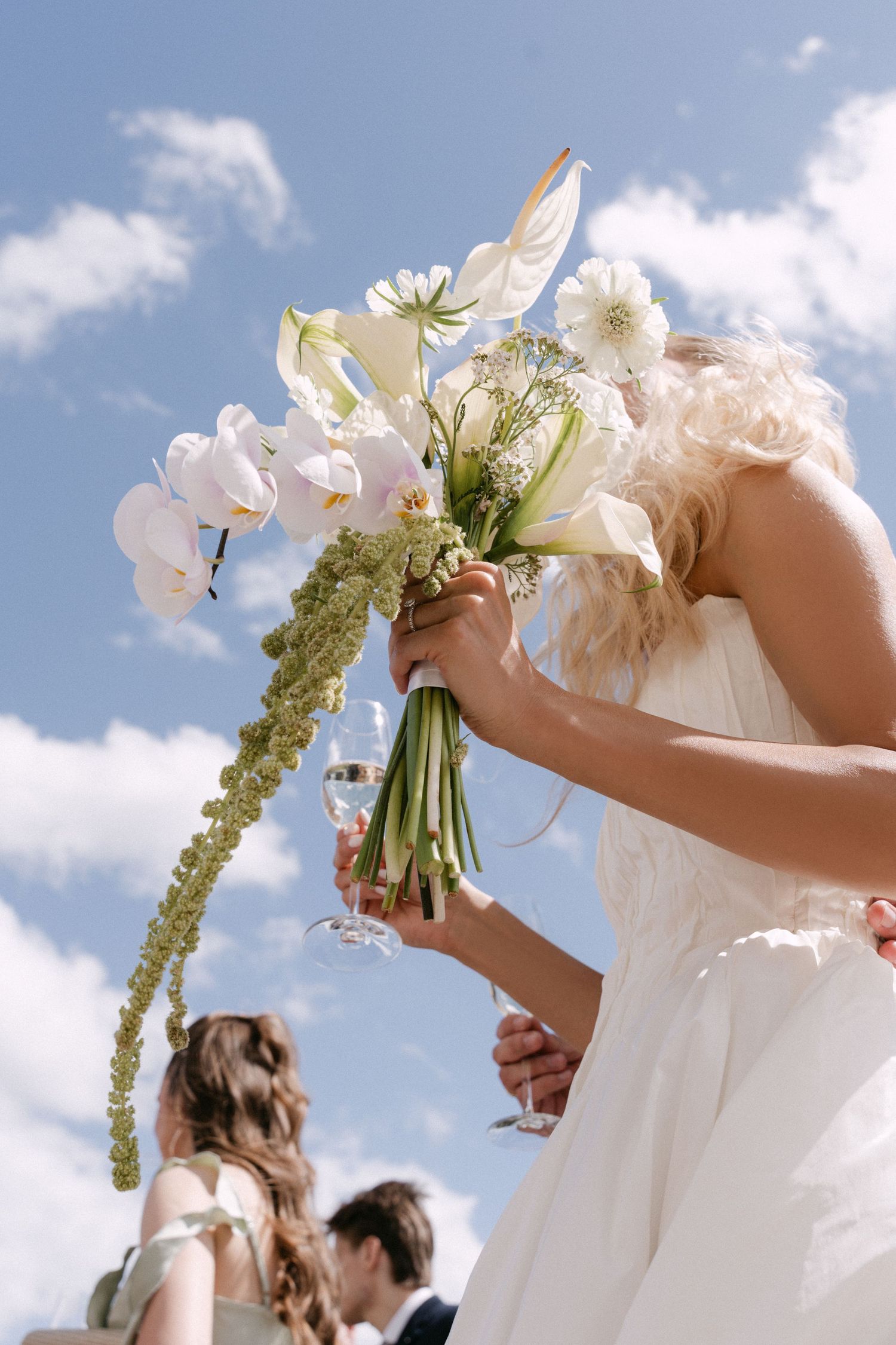 Bride holding a white wedding bouquet of orchids and calla lilies against a bright blue sky.