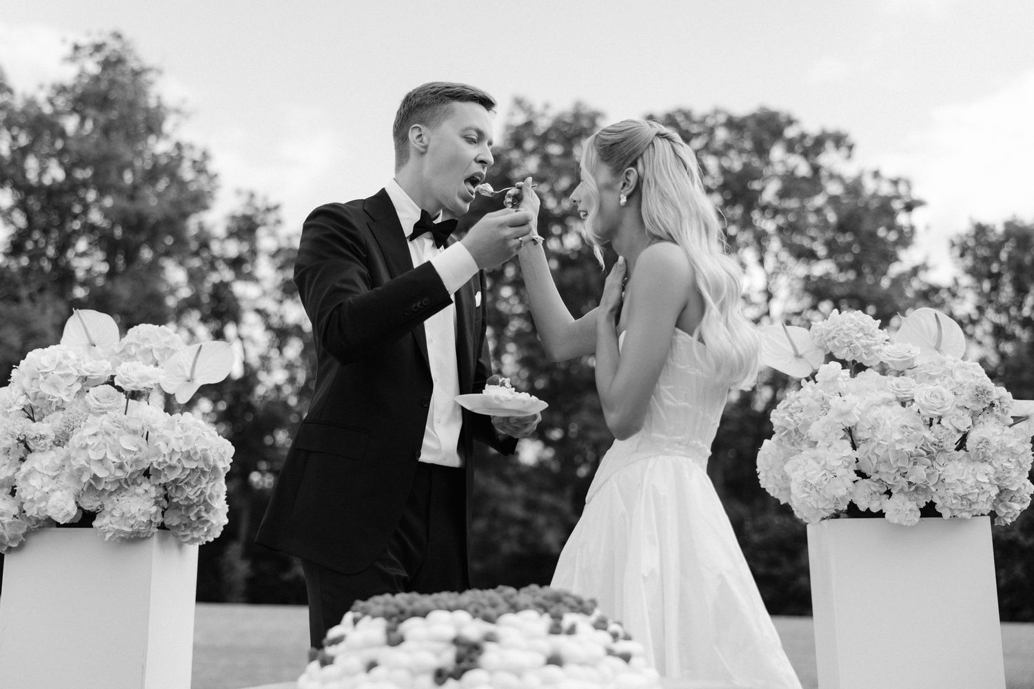 Groom feeds bride a piece of wedding cake outdoors surrounded by white hydrangea floral arrangements.