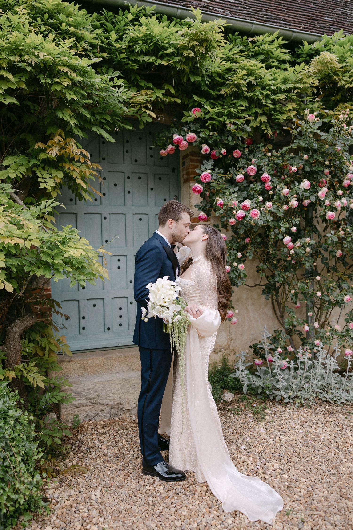 Bride and groom share a kiss near a rose-covered wall with a teal door on their wedding day.