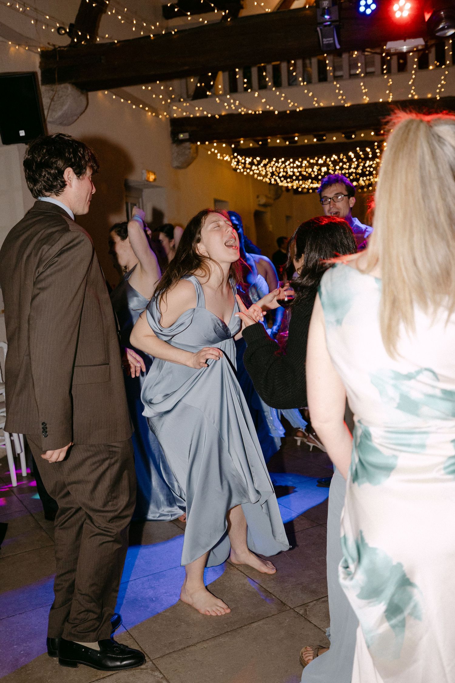 Guest in blue dress dancing barefoot energetically at lively wedding reception dance floor.