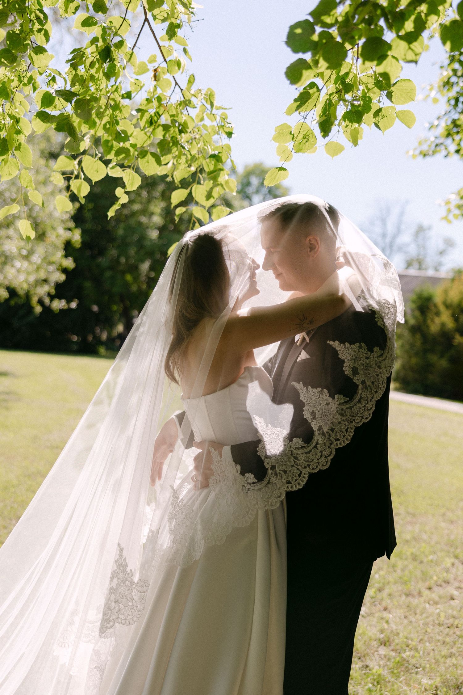 Bride and groom sharing a romantic kiss under lush green tree branches outdoors on their wedding day.