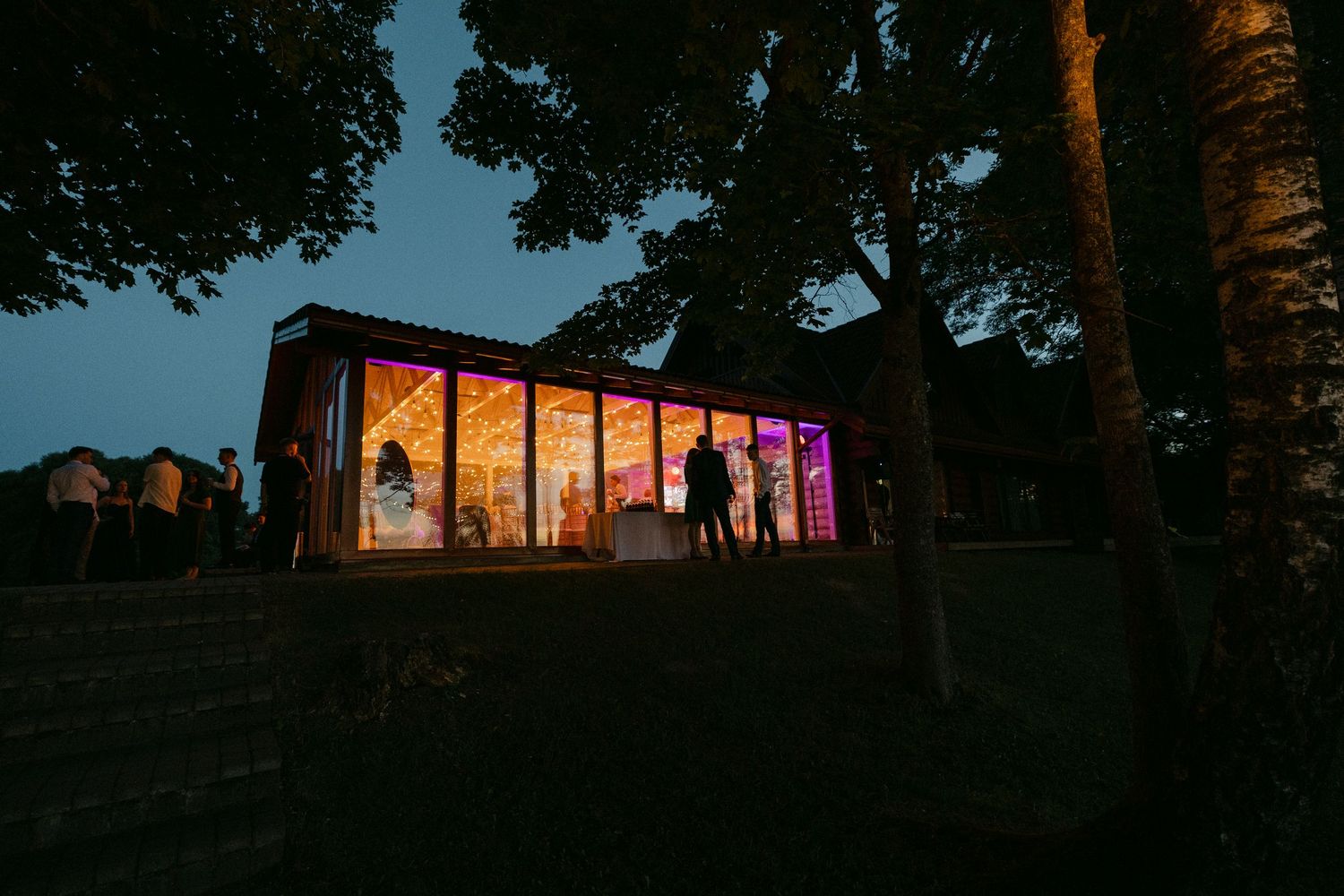 Glowing glass venue building illuminated with colorful lights at dusk surrounded by silhouetted trees.