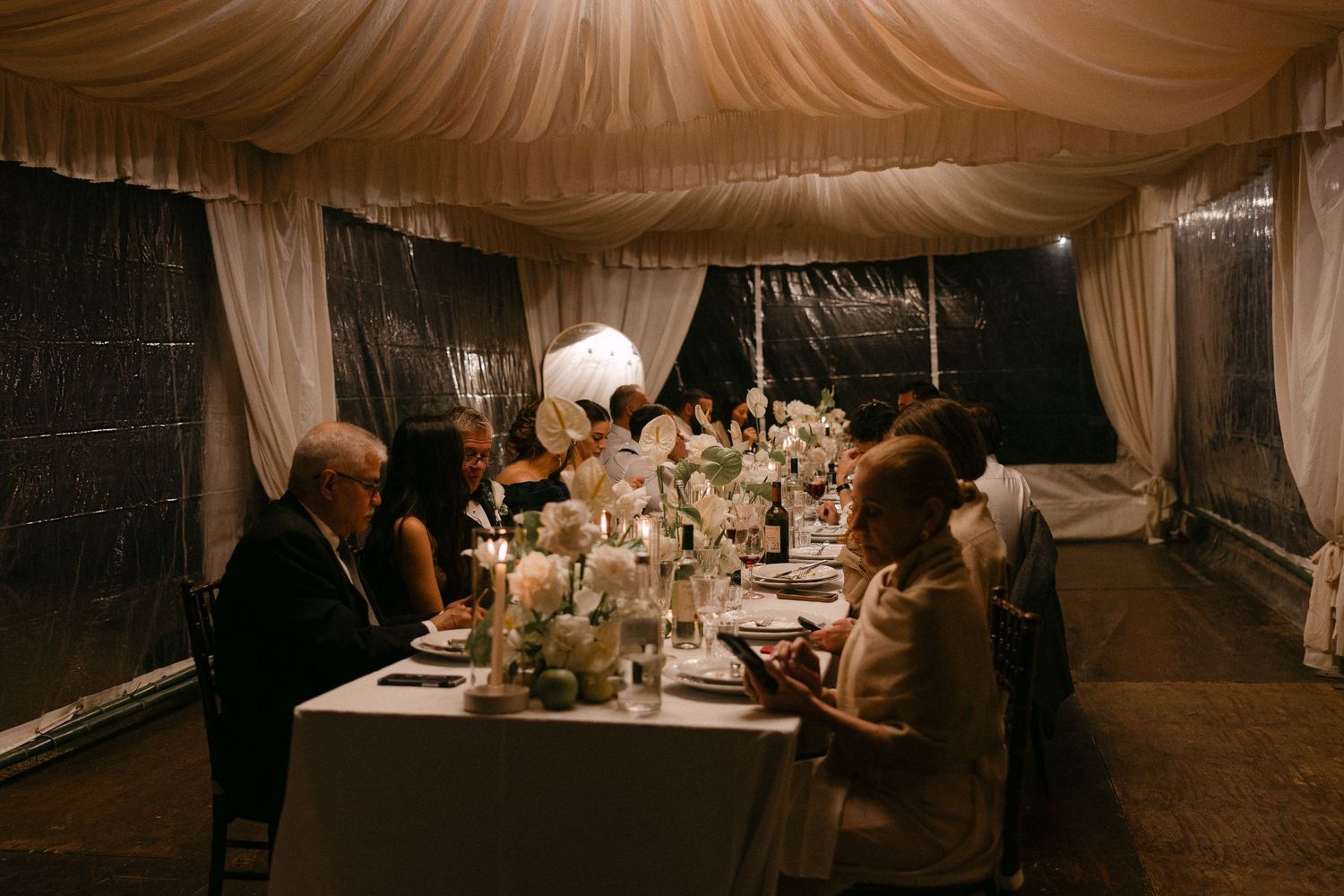 Wedding guests dine by candlelight under draped tent with floral centerpieces at elegant reception.