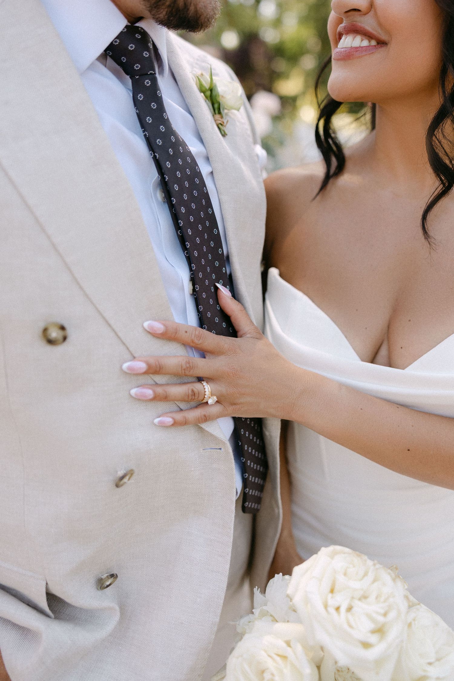 Close-up of bride in white gown resting hand on groom's linen suit jacket holding white roses.