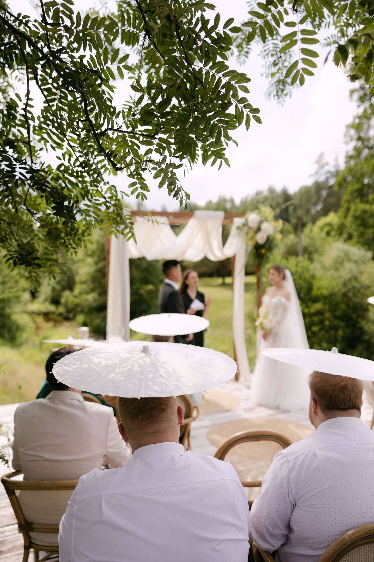 Guests seated outdoors watch a wedding ceremony under a floral arch surrounded by lush green trees.