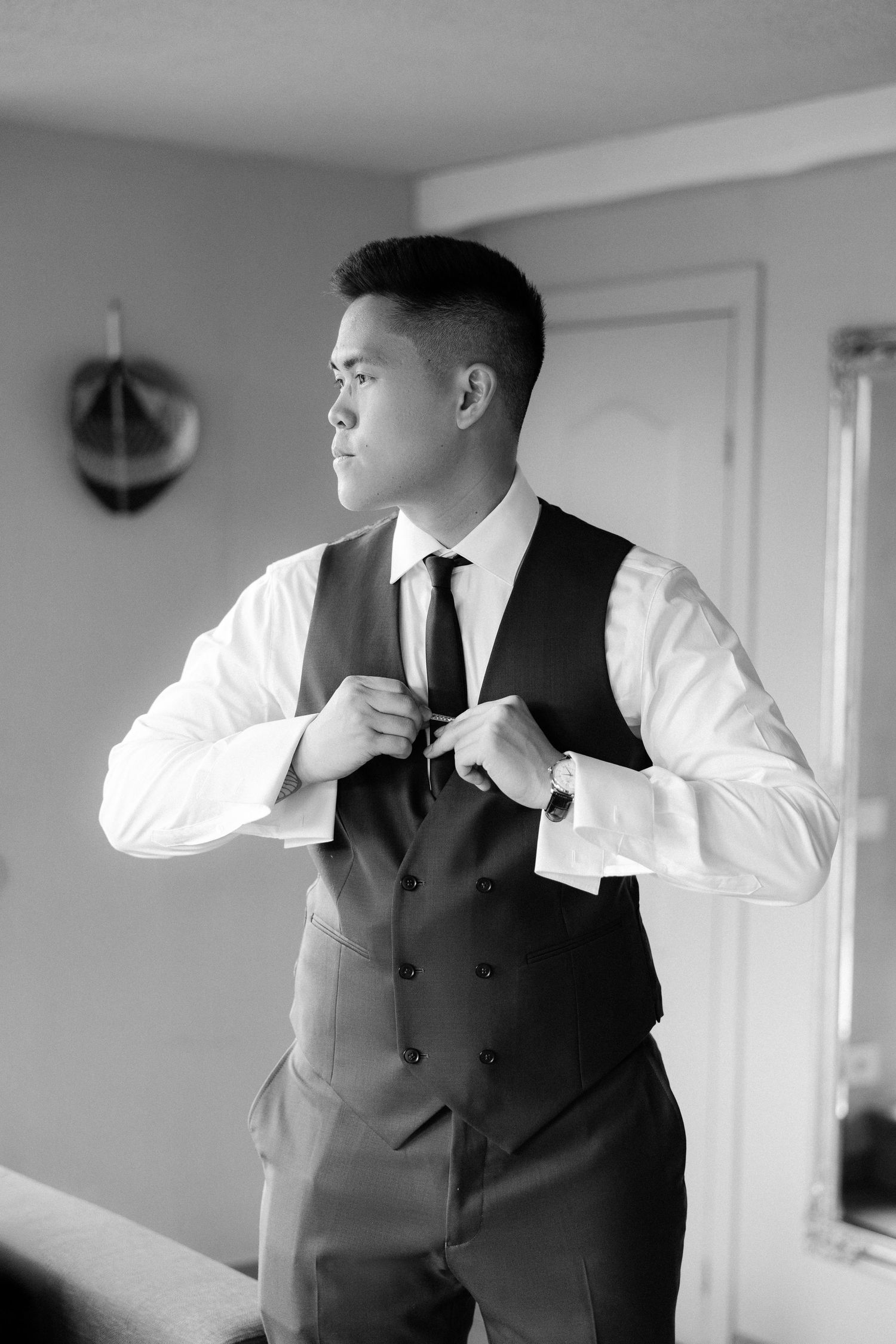 Black and white photo of a groom adjusting his tie while wearing a vest and dress shirt before his wedding.