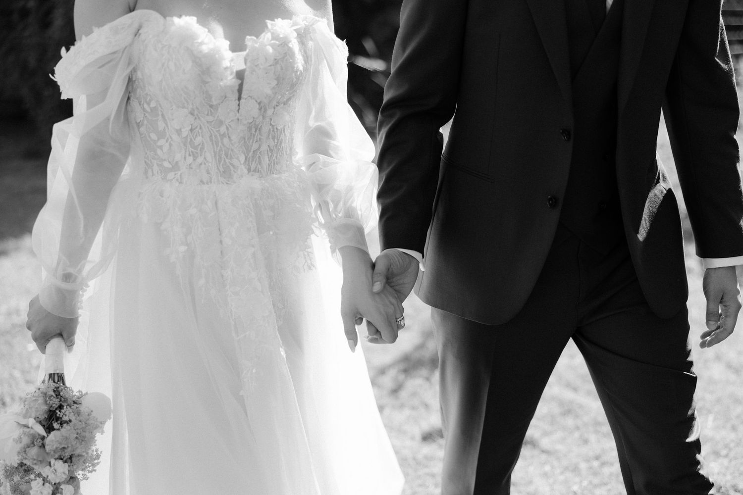 Black and white photo of bride in lace gown holding bouquet walking hand in hand with groom.