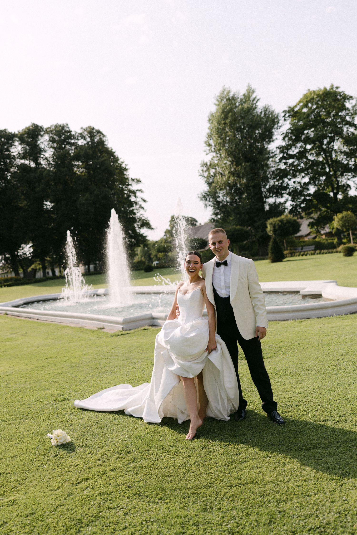Bride lifting white gown and groom in ivory tuxedo pose by fountain in lush garden.