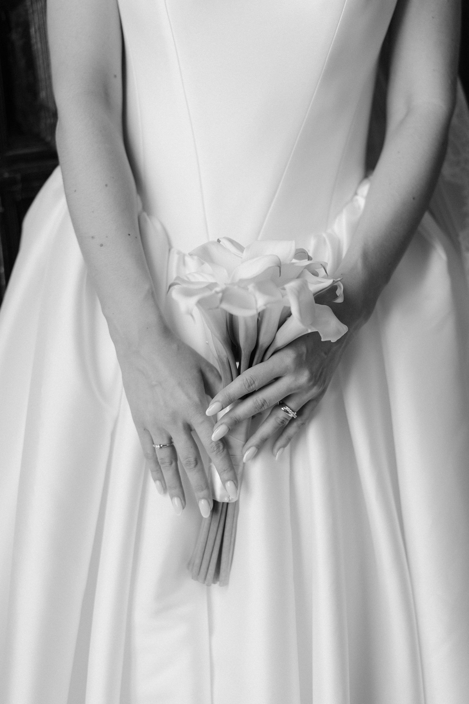 Bride in white gown holding elegant calla lily bouquet, close-up of hands with wedding ring, black and white photo.