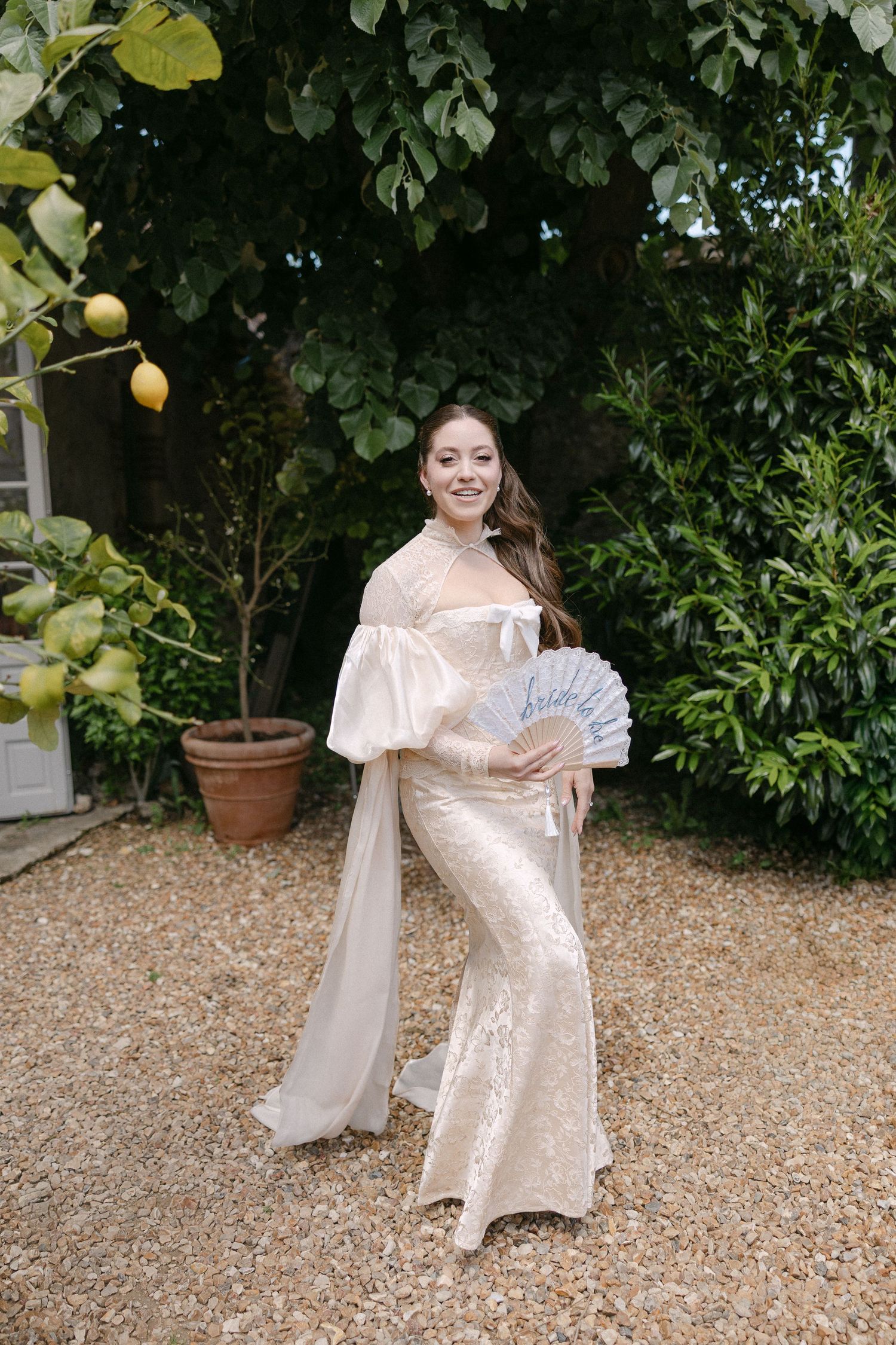 Bride in ivory satin gown holding a decorative fan standing beside a lemon tree in a garden.
