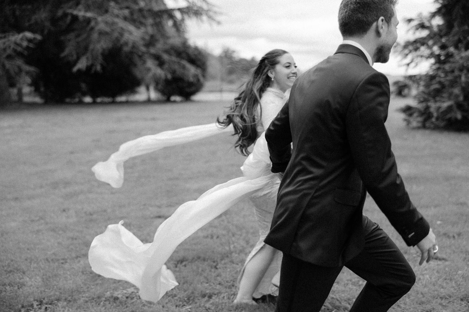 Black and white photo of bride and groom walking across lawn, veil flowing in the wind.