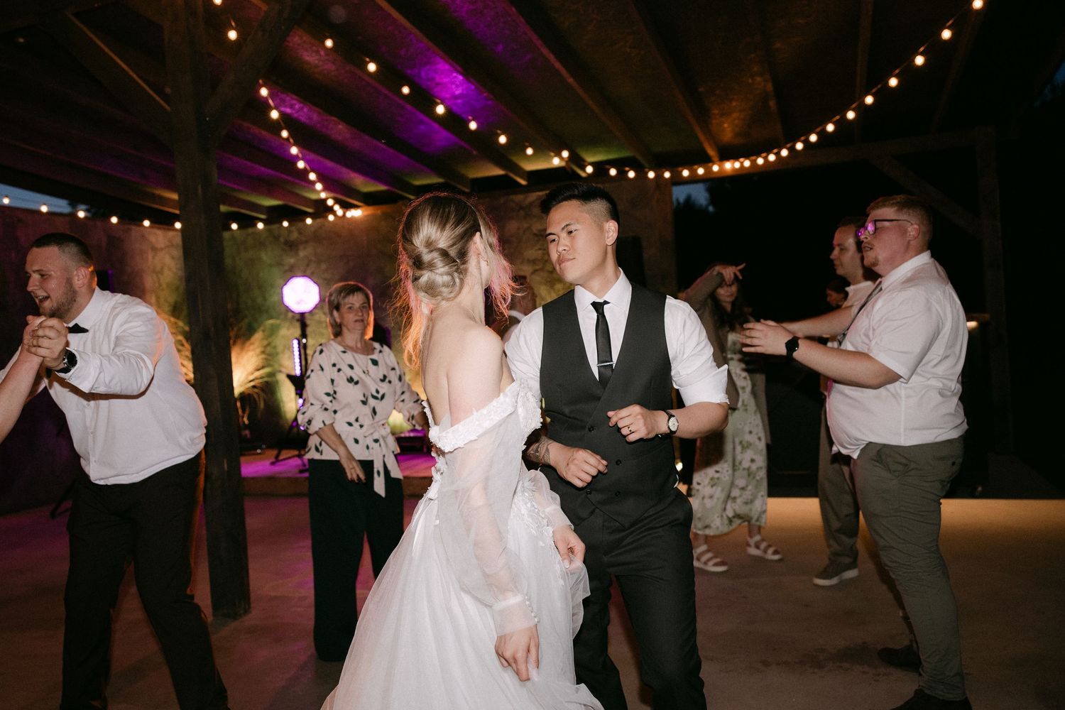 Bride and groom share first dance surrounded by guests under string lights at indoor wedding reception.