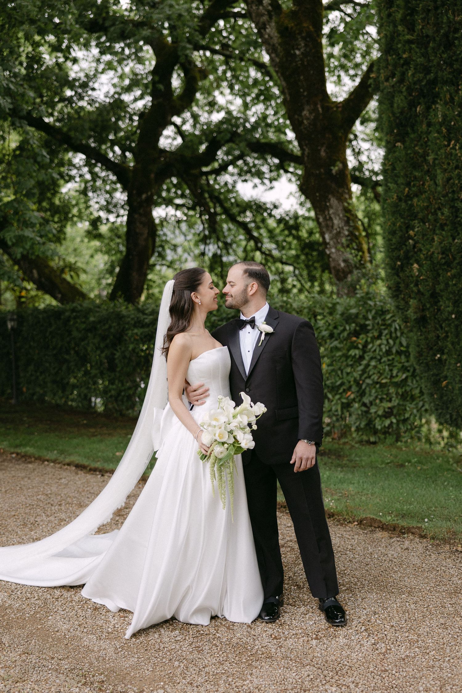 Bride and groom share a romantic moment on a gravel path surrounded by lush green trees.