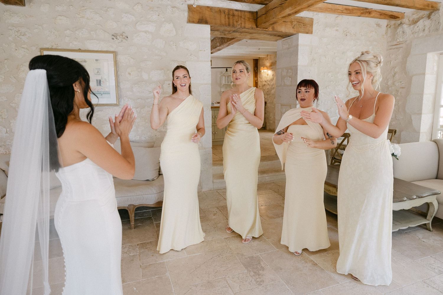 Bride reveals wedding dress to bridesmaids in cream gowns, all reacting with joy in a rustic stone venue.