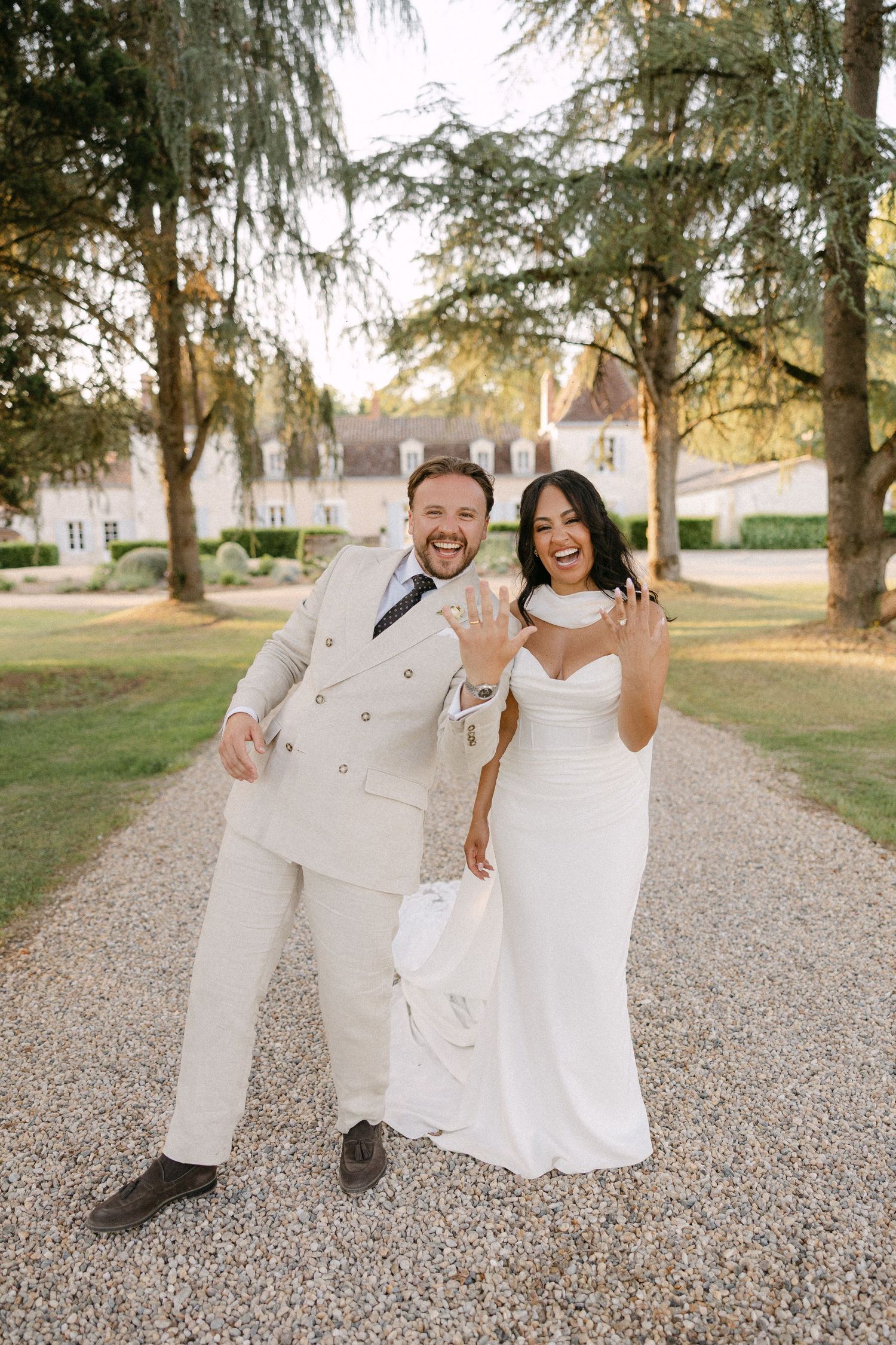 Joyful newlyweds showing wedding rings while laughing on gravel driveway of French chateau.