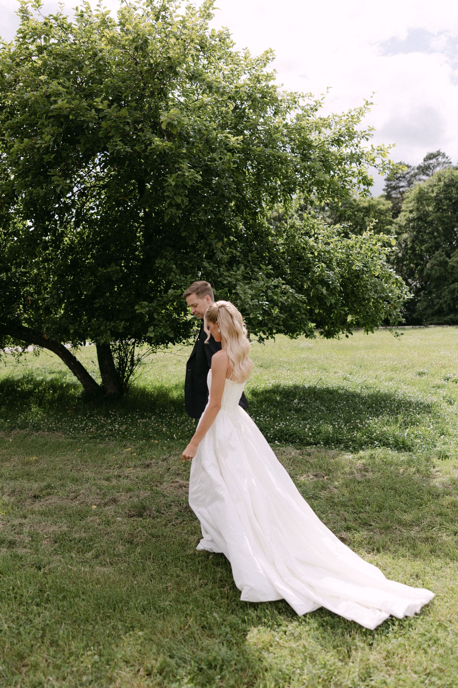 Bride in a flowing white gown and groom in black suit walking together in a lush green garden.