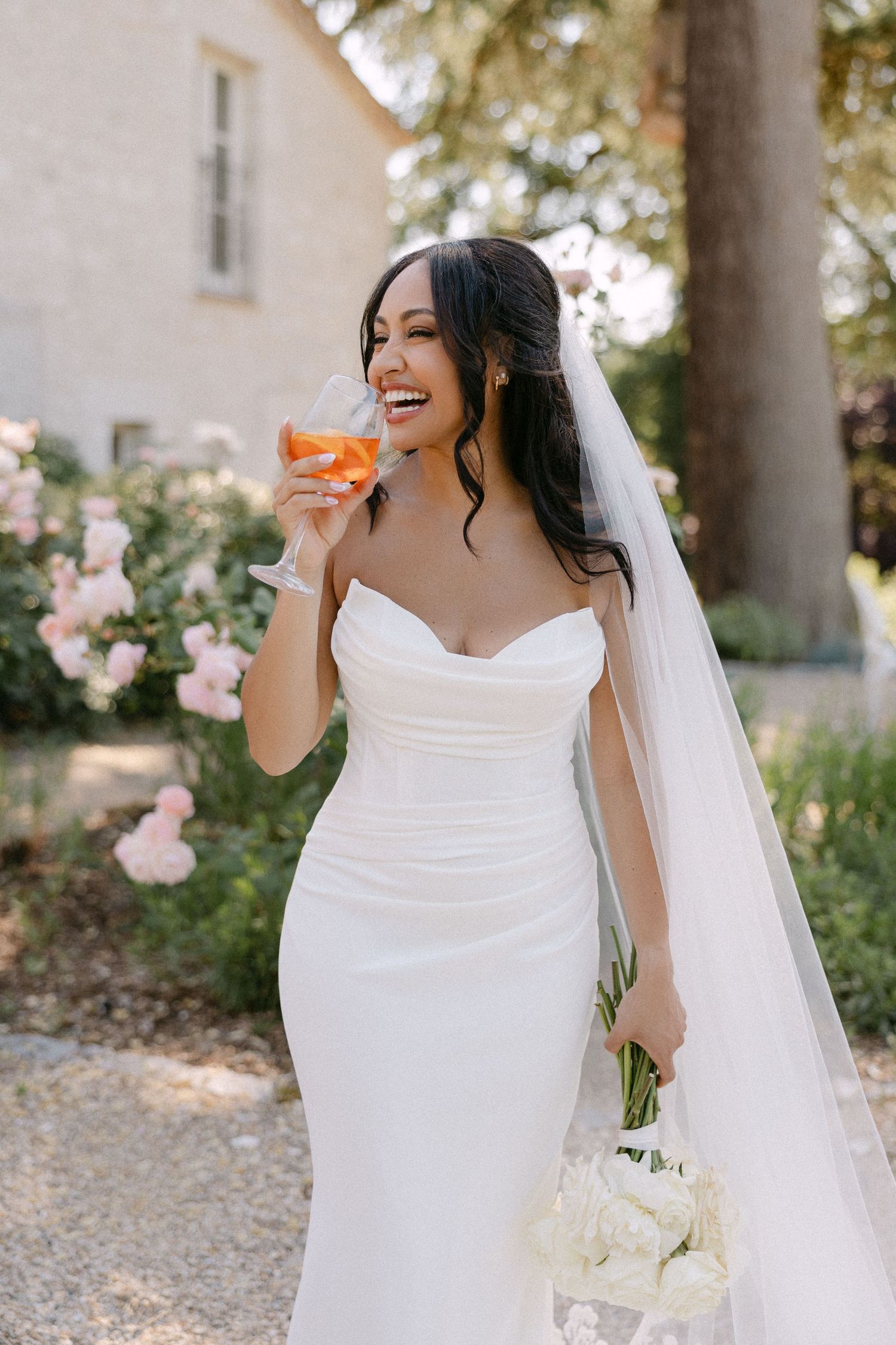 Radiant bride in strapless white gown and veil holds bouquet and champagne glass in garden setting.