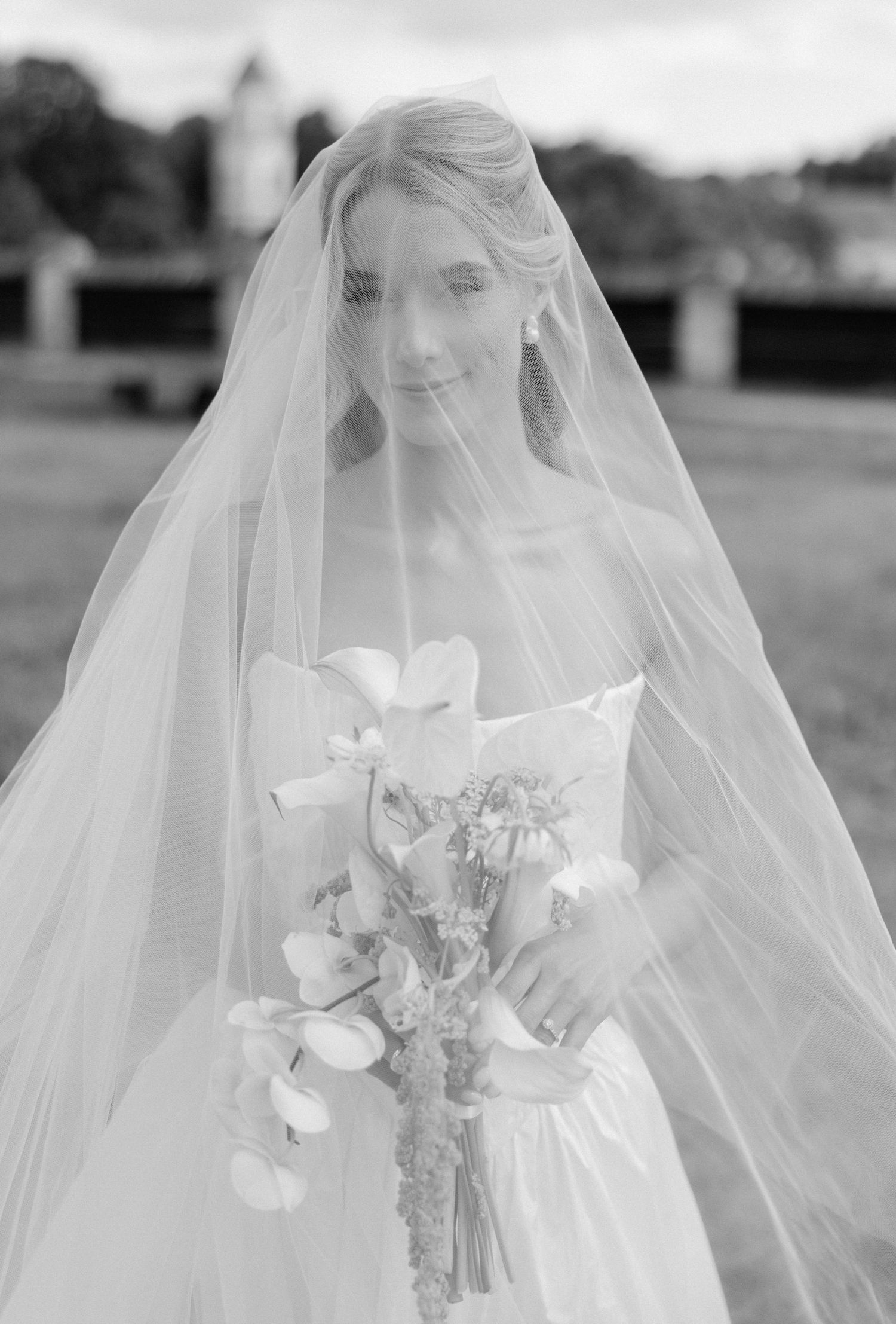 Black and white portrait of a bride wearing a flowing veil holding a floral bouquet outdoors.