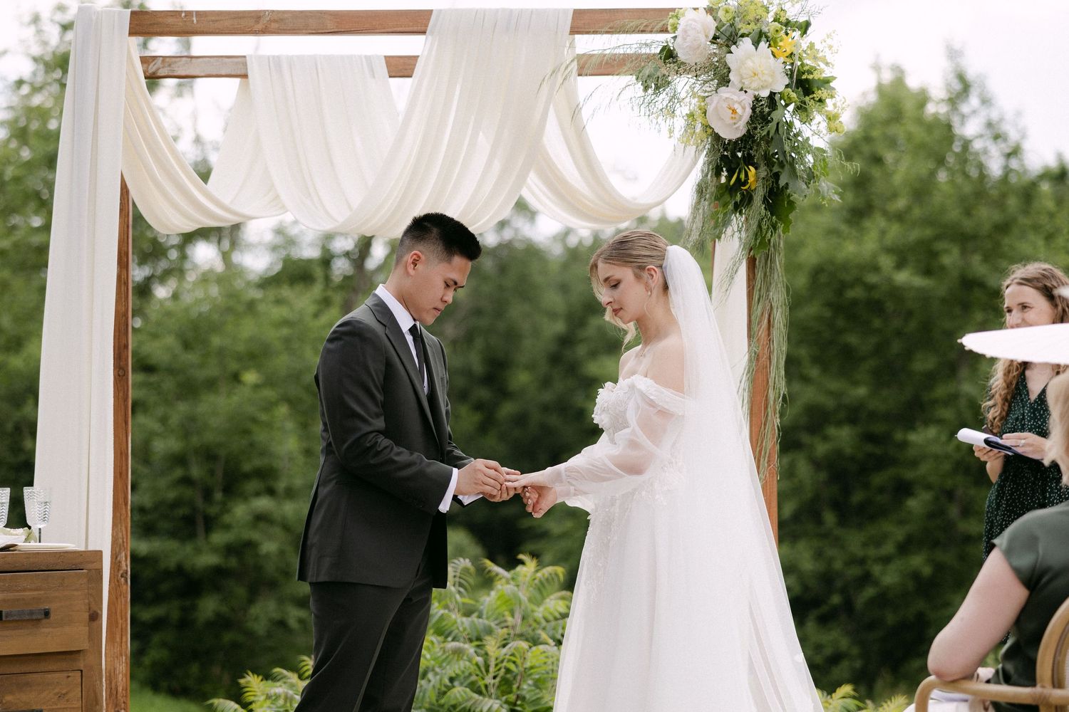 Groom placing ring on bride's finger during outdoor wedding ceremony under a wooden arch with white draping and white roses.