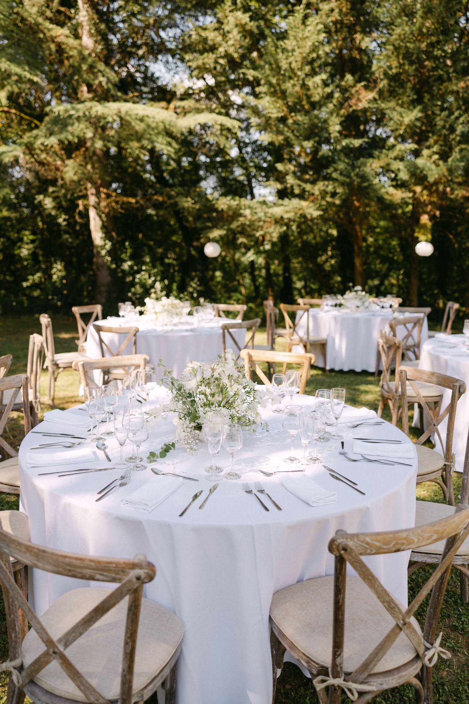Elegant outdoor wedding reception with white round tables, wooden cross-back chairs, and floral centerpieces set in a lush garden.