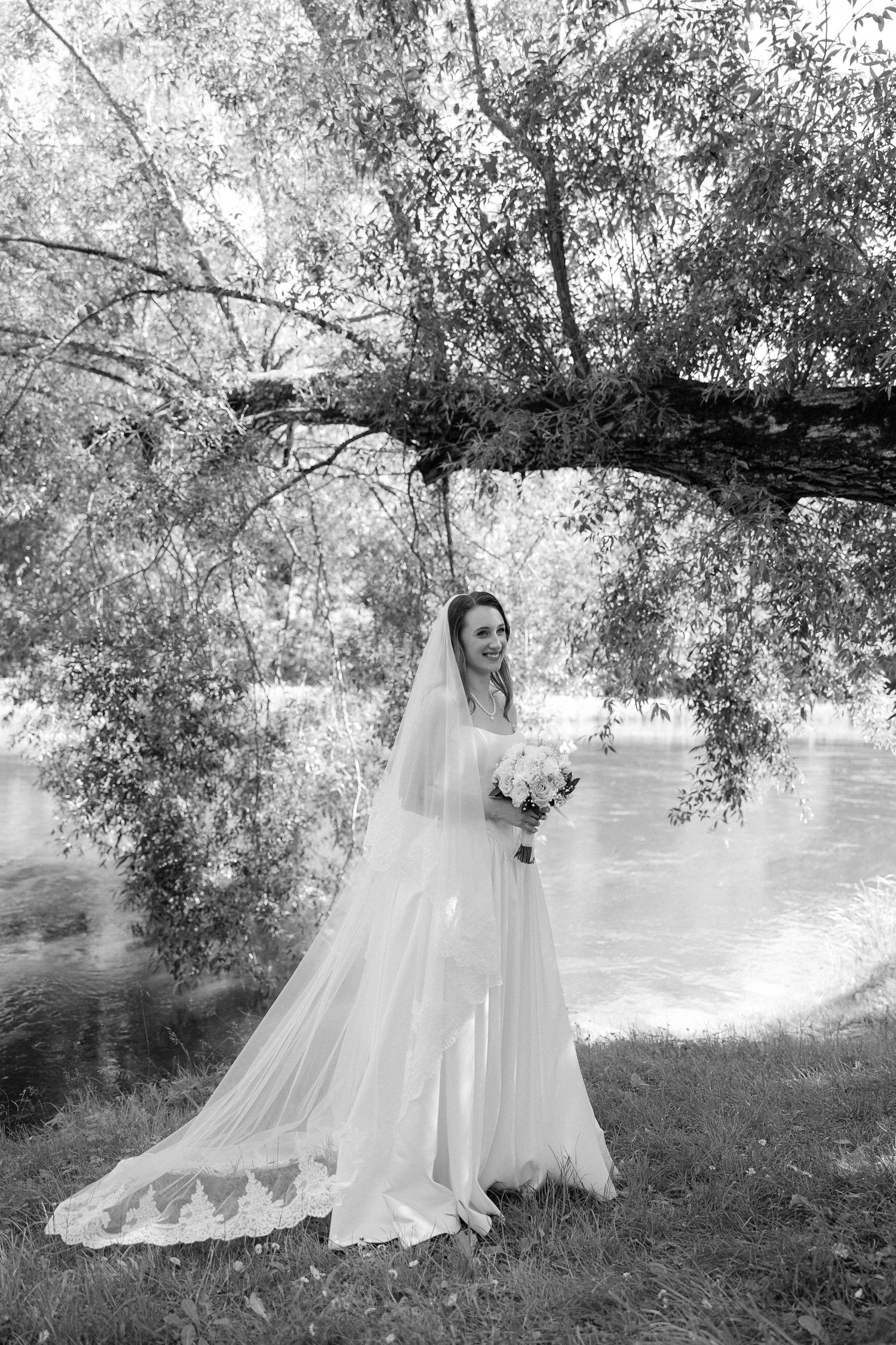 Black and white portrait of bride in lace-trimmed wedding gown and veil standing by riverside tree.