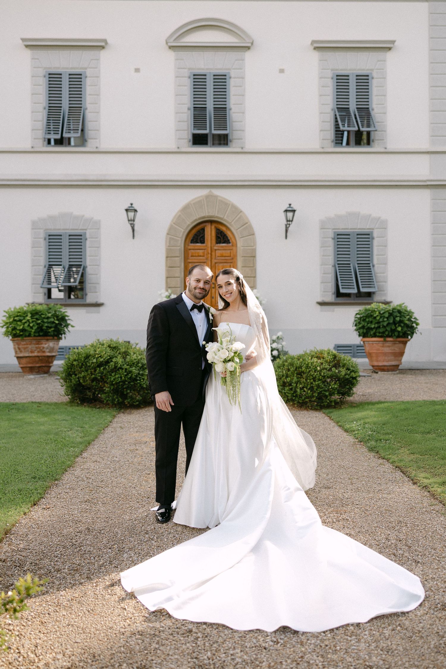 Smiling bride and groom pose together in front of elegant white villa with green shutters on wedding day.